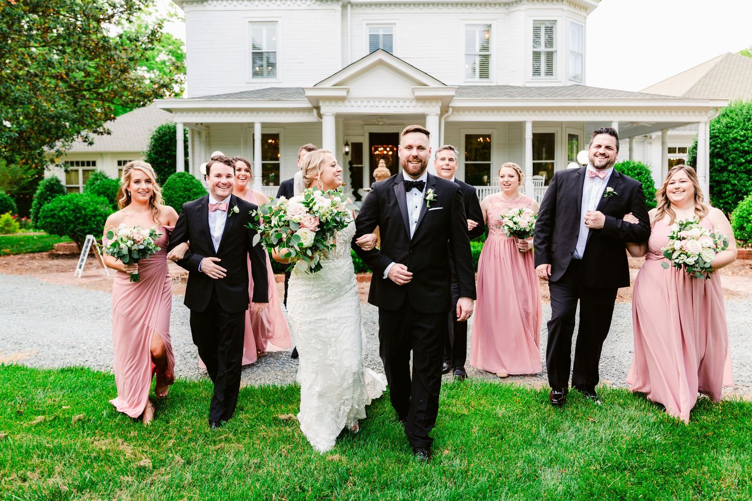 Wedding party in pink bridesmaid dresses and black tuxedos walk on grass in front of white house.
