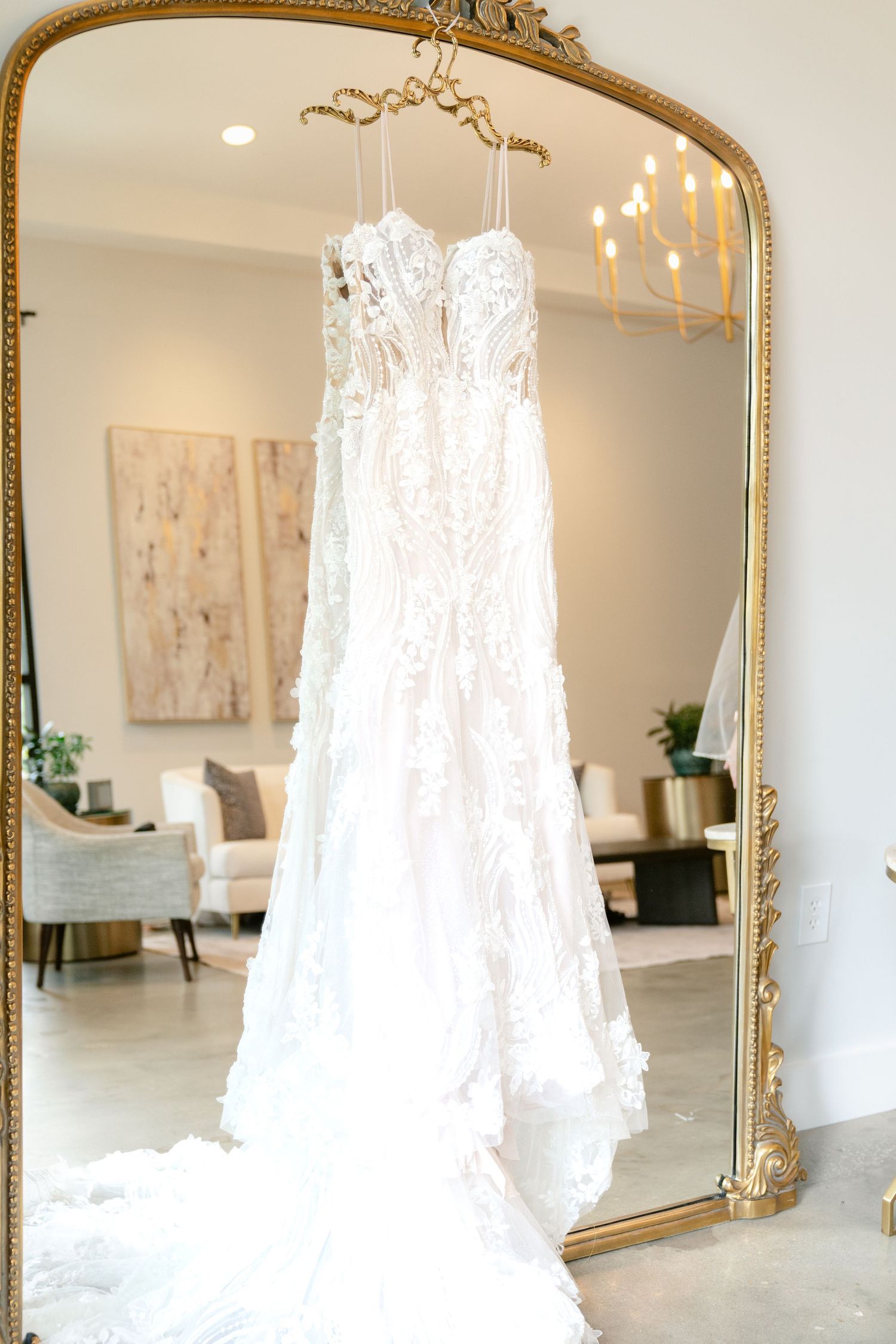 A delicate white lace wedding dress hangs from a gold-framed mirror in an elegant room with a chandelier.