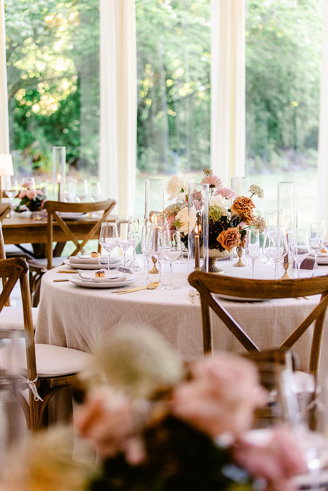 Light-filled reception venue with cross-back chairs and pink floral centerpieces on round tables.
