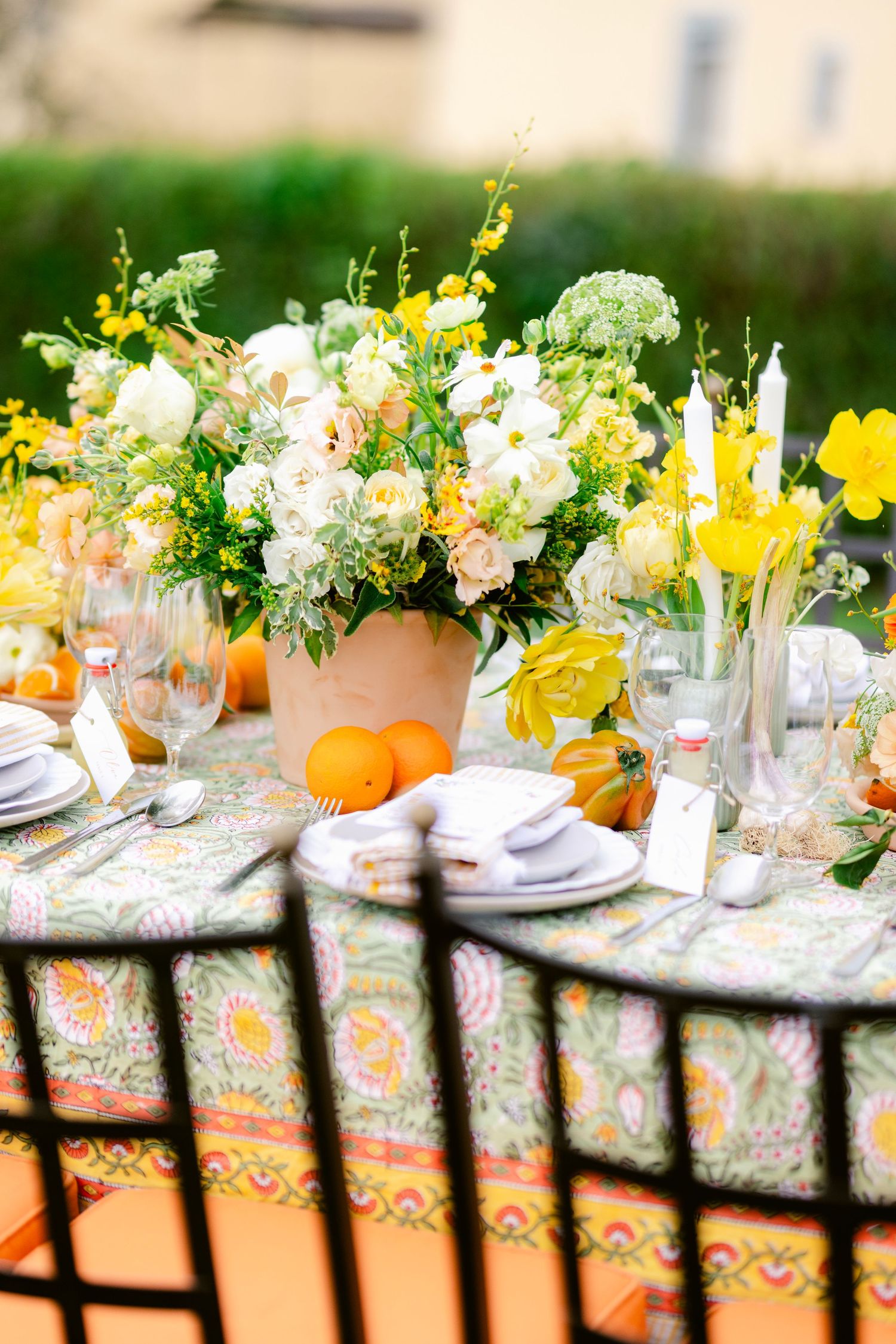 Elegant outdoor table setting with colorful floral centerpieces, orange accents, and crystal glassware on a patterned tablecloth.