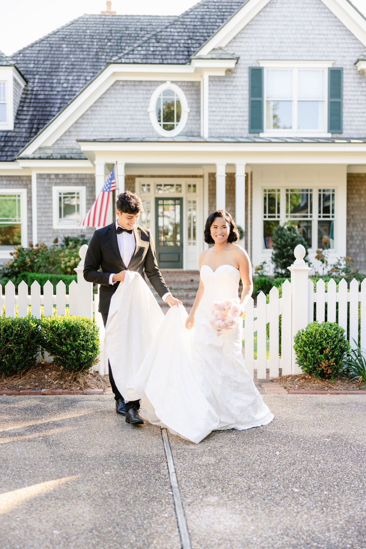 A wedding couple walks along a gravel path in front of white picket fences and charming cottage-style buildings.