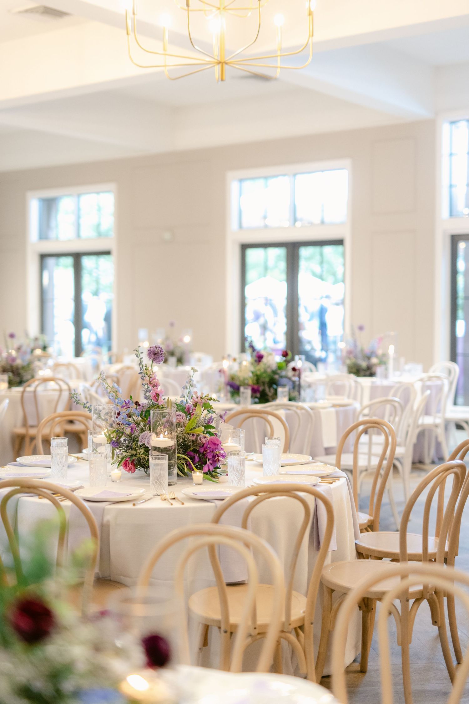 Elegant wedding reception venue with white tables, bentwood chairs and colorful floral centerpieces in a bright room.