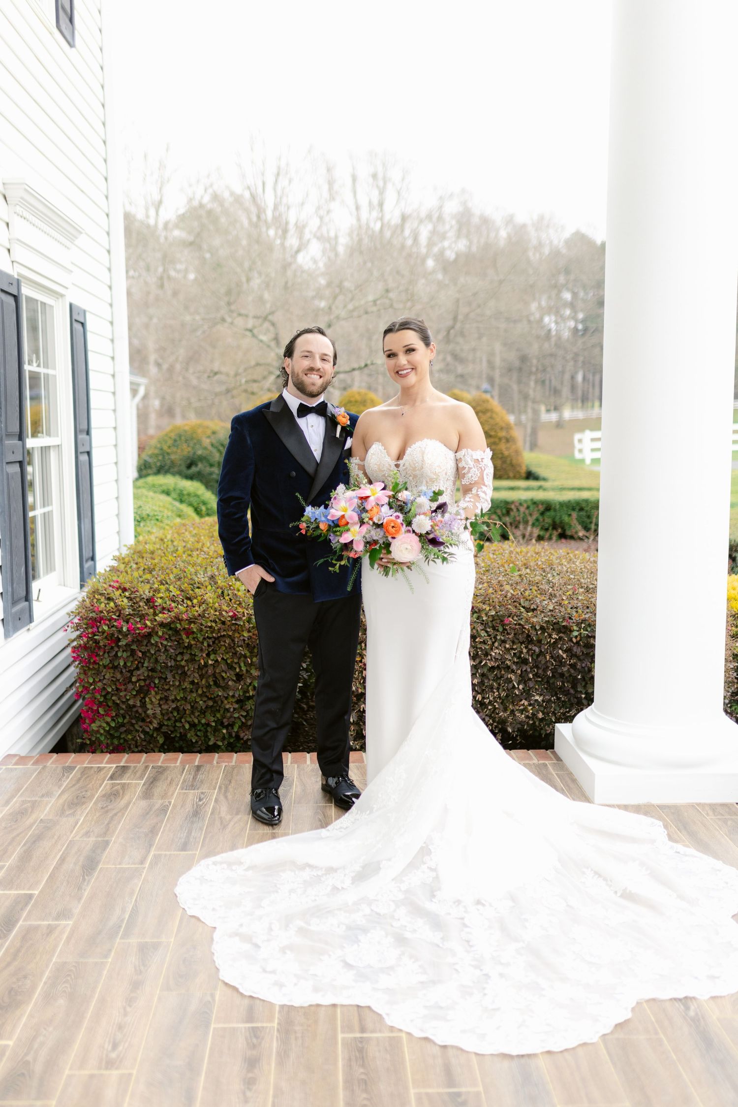 A couple in wedding attire poses together on a covered porch with white columns and manicured shrubs in the background.