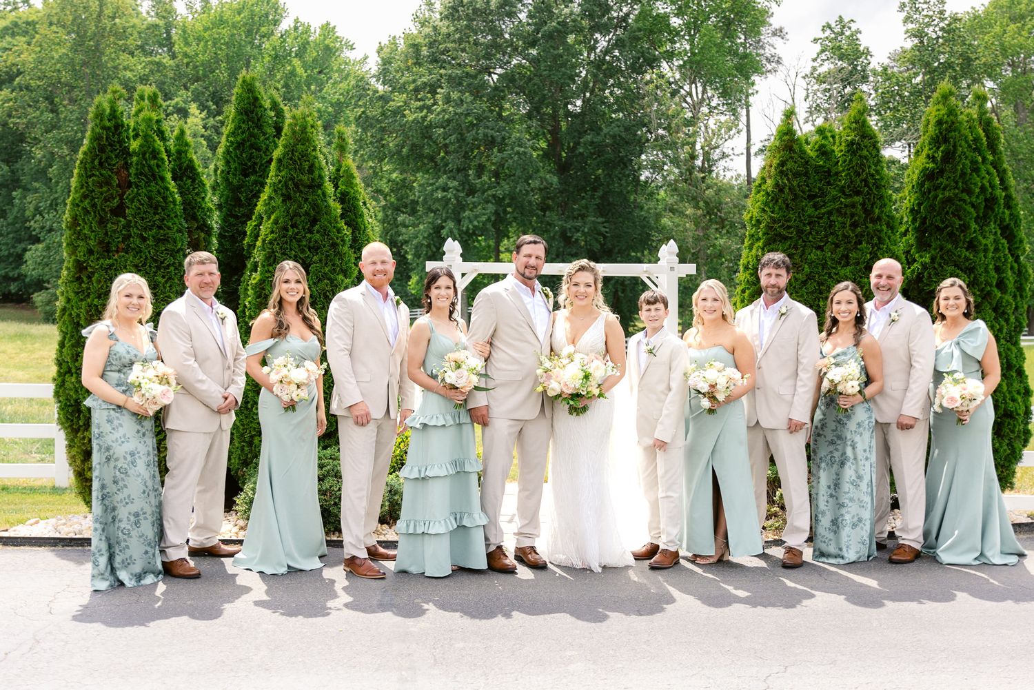 Wedding party in pastel mint and beige attire poses outdoors with tall evergreen trees in background.