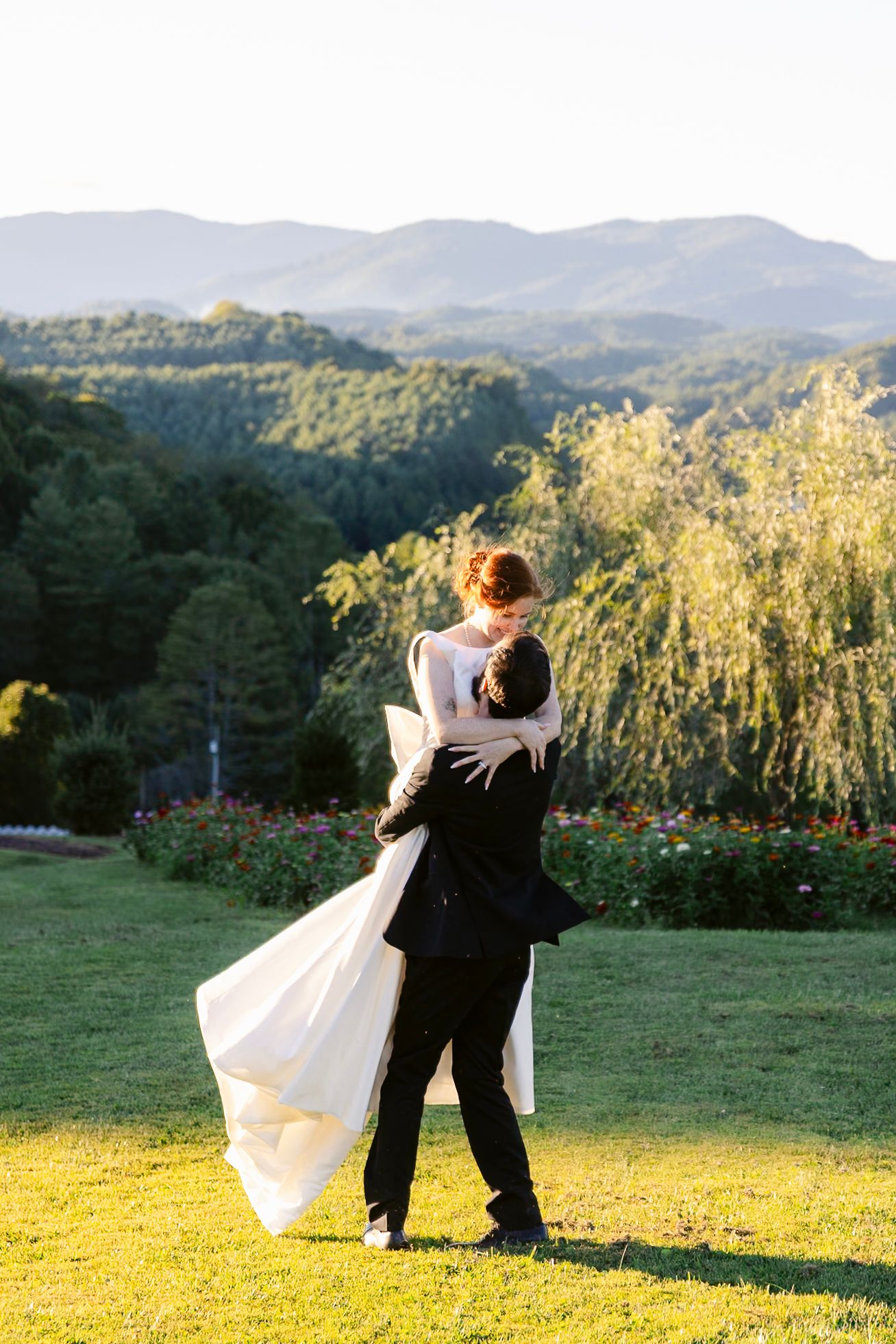 A couple in wedding attire embraces while standing on a grassy hilltop with mountains and golden sunlight in the background.