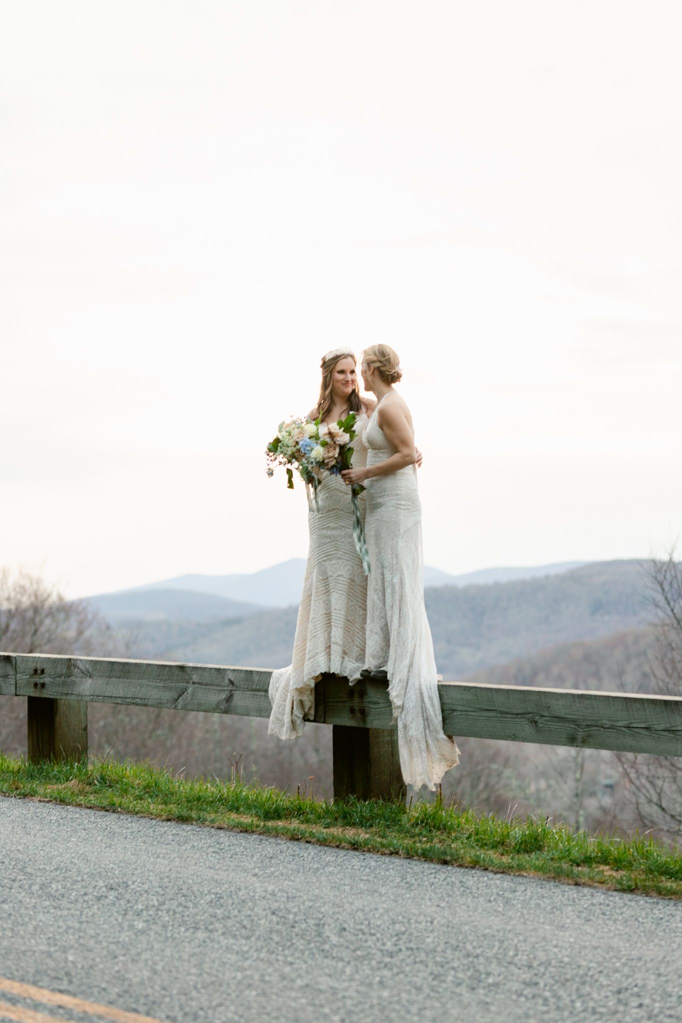 Two people in white dresses embrace while standing against a wooden railing with mountain views.