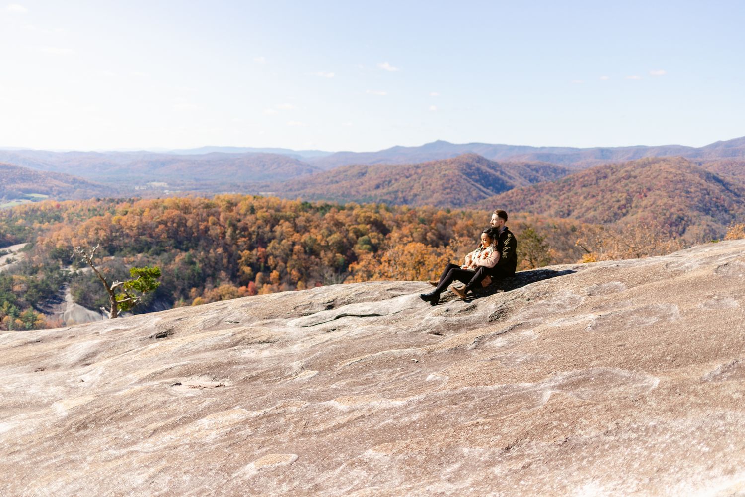 Two people sit on a rocky mountain overlook surrounded by colorful fall foliage in the valley below.