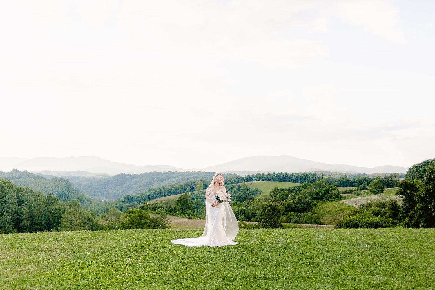 A panoramic view of a rolling green hillside with distant mountains and a figure in white standing in the meadow.
