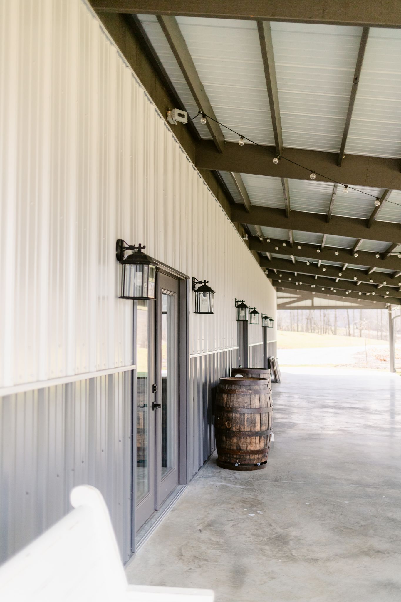 Long covered walkway with concrete floor and white metal walls features mounted light fixtures and a rustic wooden barrel.