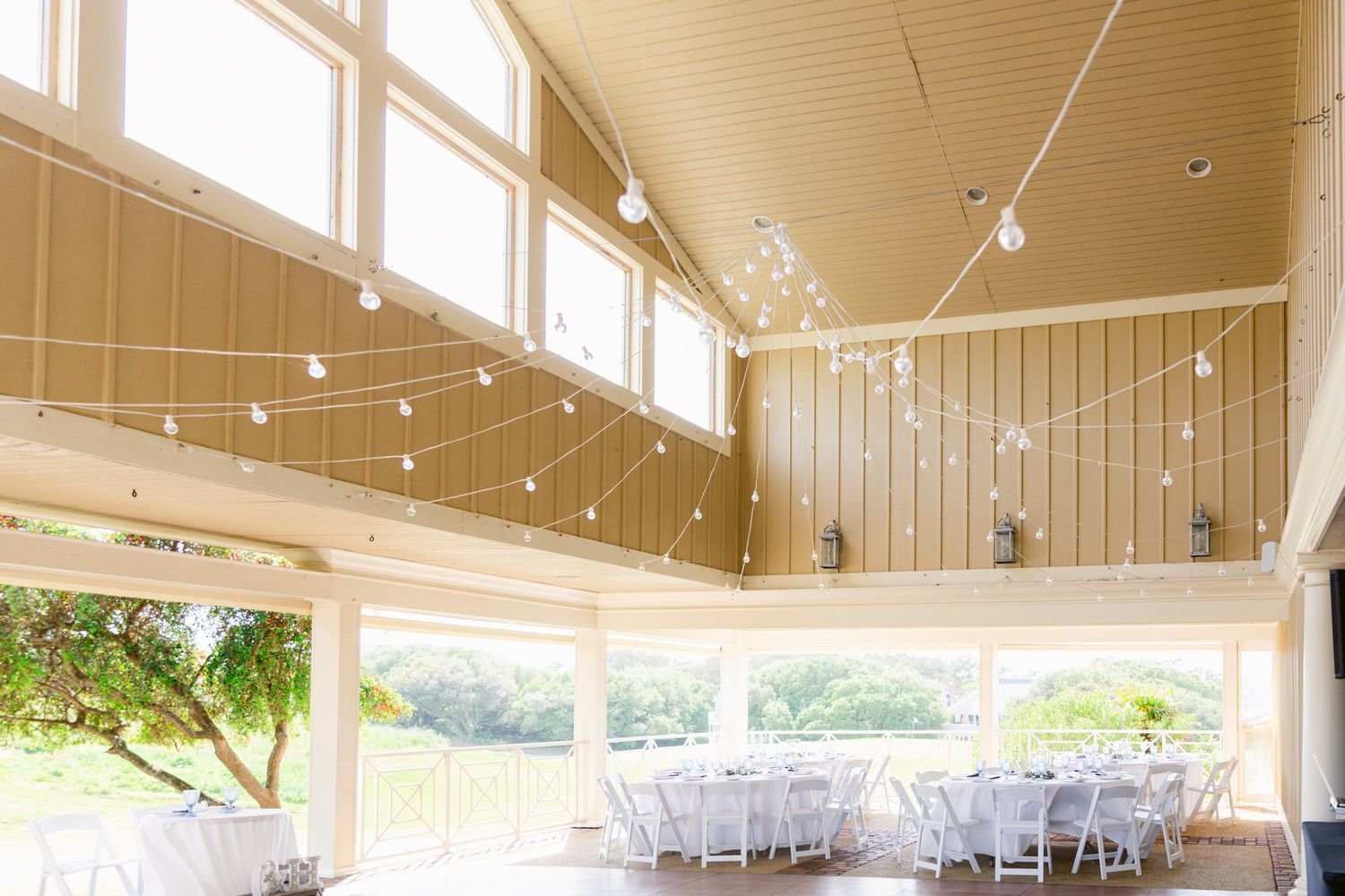 A spacious event venue interior with wooden beams, string lights, and white chairs set up for a celebration.