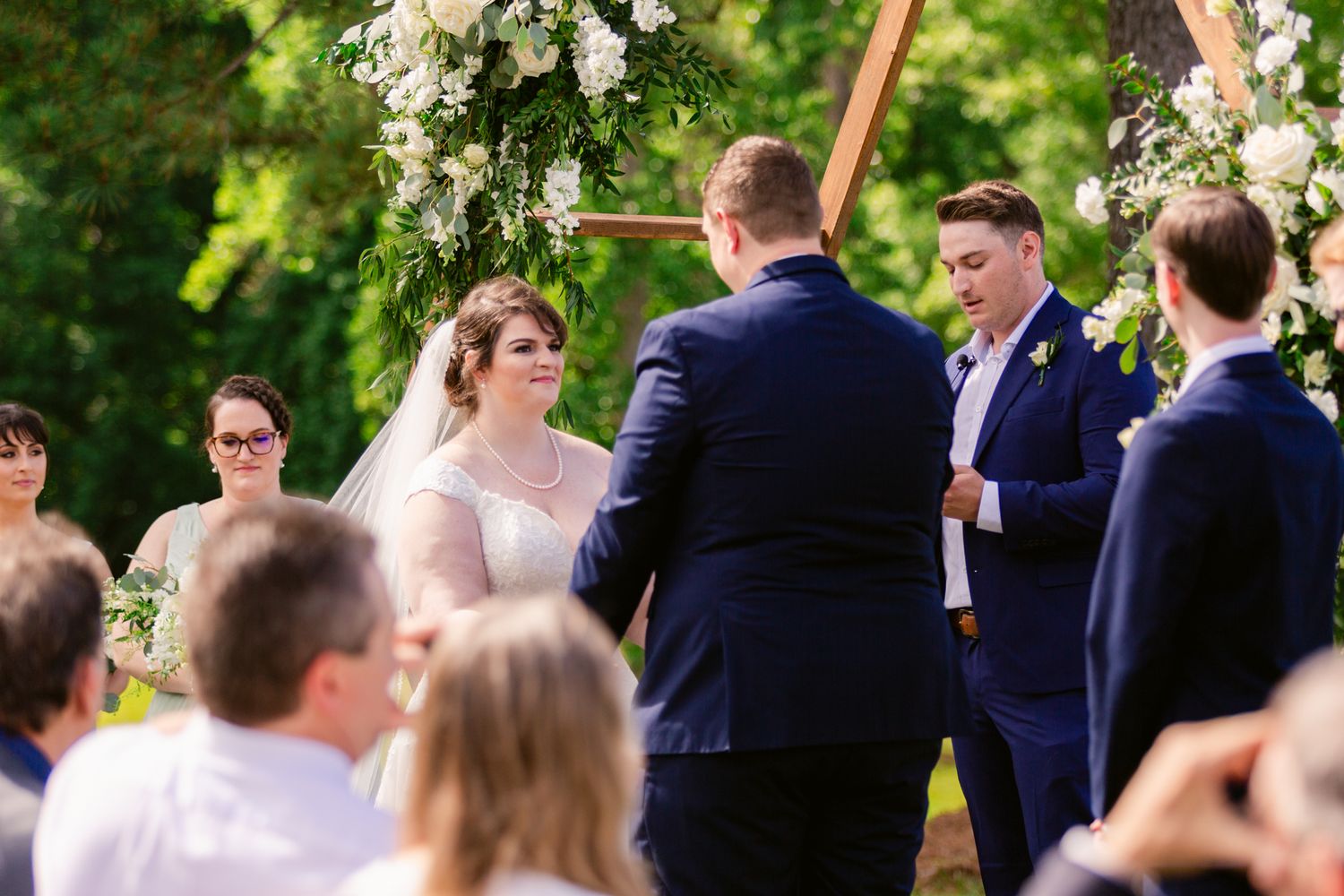 Wedding guests gather outdoors under white floral decorations and greenery during a garden ceremony.
