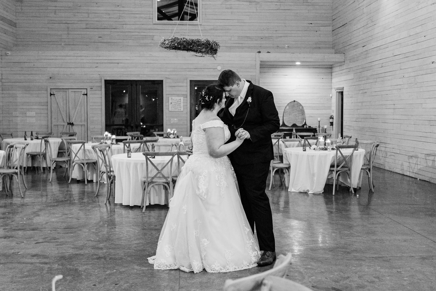 A couple shares their first dance in a rustic white-walled reception venue with round tables and cross-back chairs.