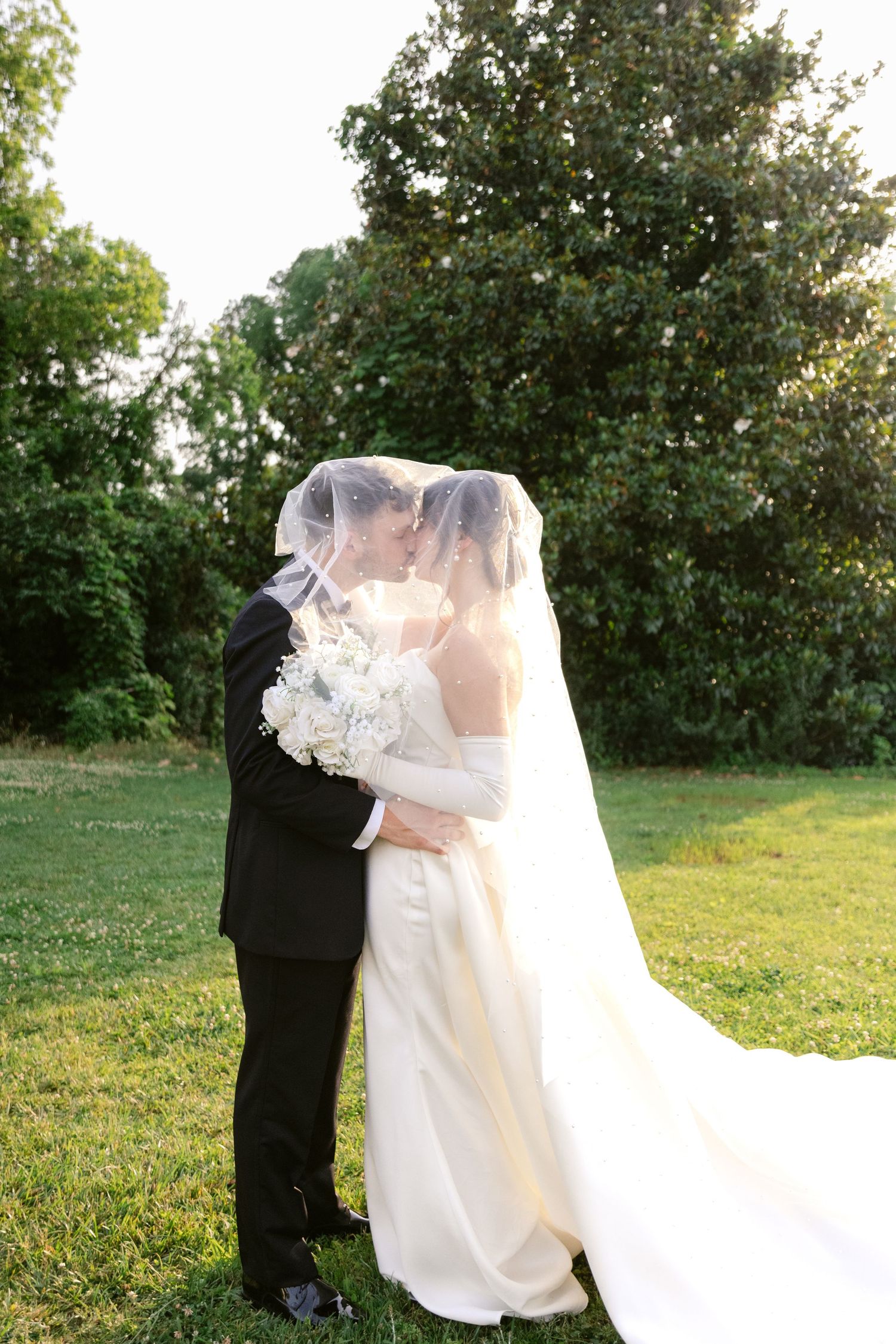 A romantic wedding couple embraces outdoors in a garden setting with a flowing white veil and dress at sunset.