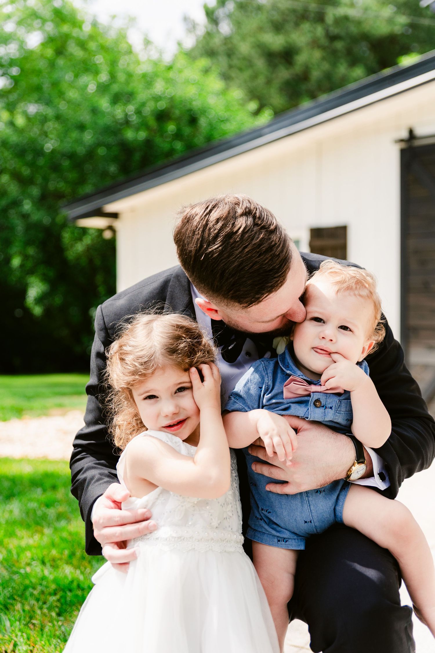 A loving family moment captured outdoors with two young children and an adult sharing affectionate embraces on a sunny day.
