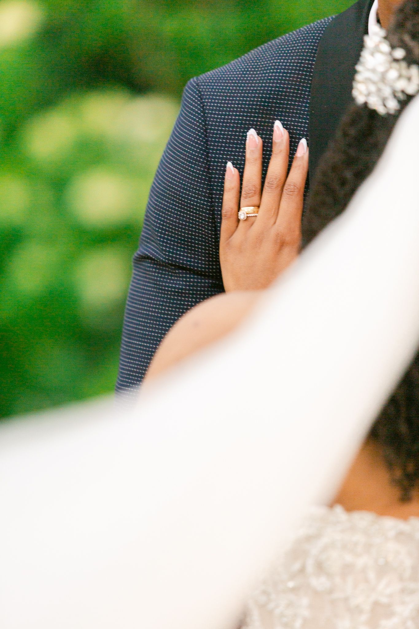 Close-up detail of a wedding ring and manicured nails against a navy suit jacket.