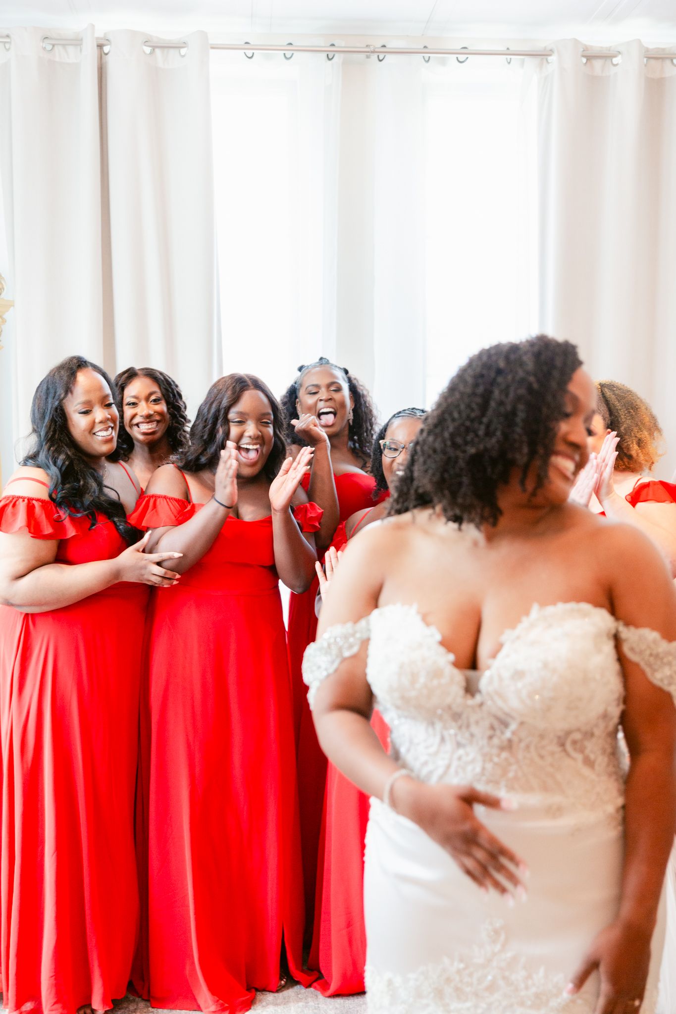 A wedding party in bright red bridesmaid dresses and white bridal gown laughing and embracing in a joyful group moment.