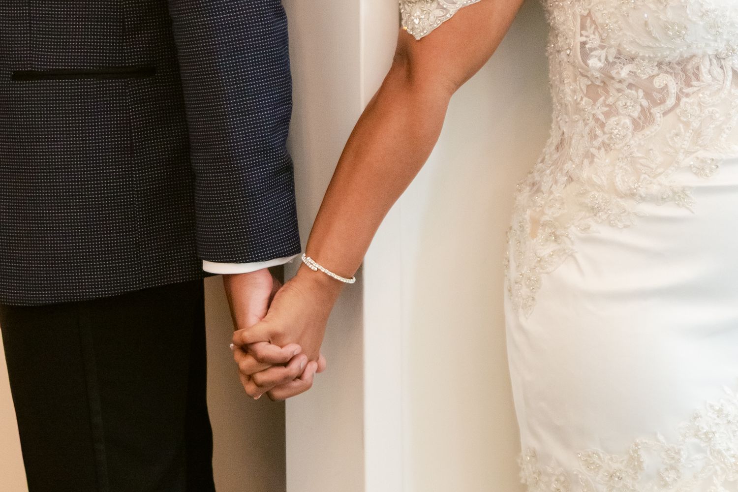 Close up of a bride and groom holding hands, showing white lace wedding dress and dark suit details.