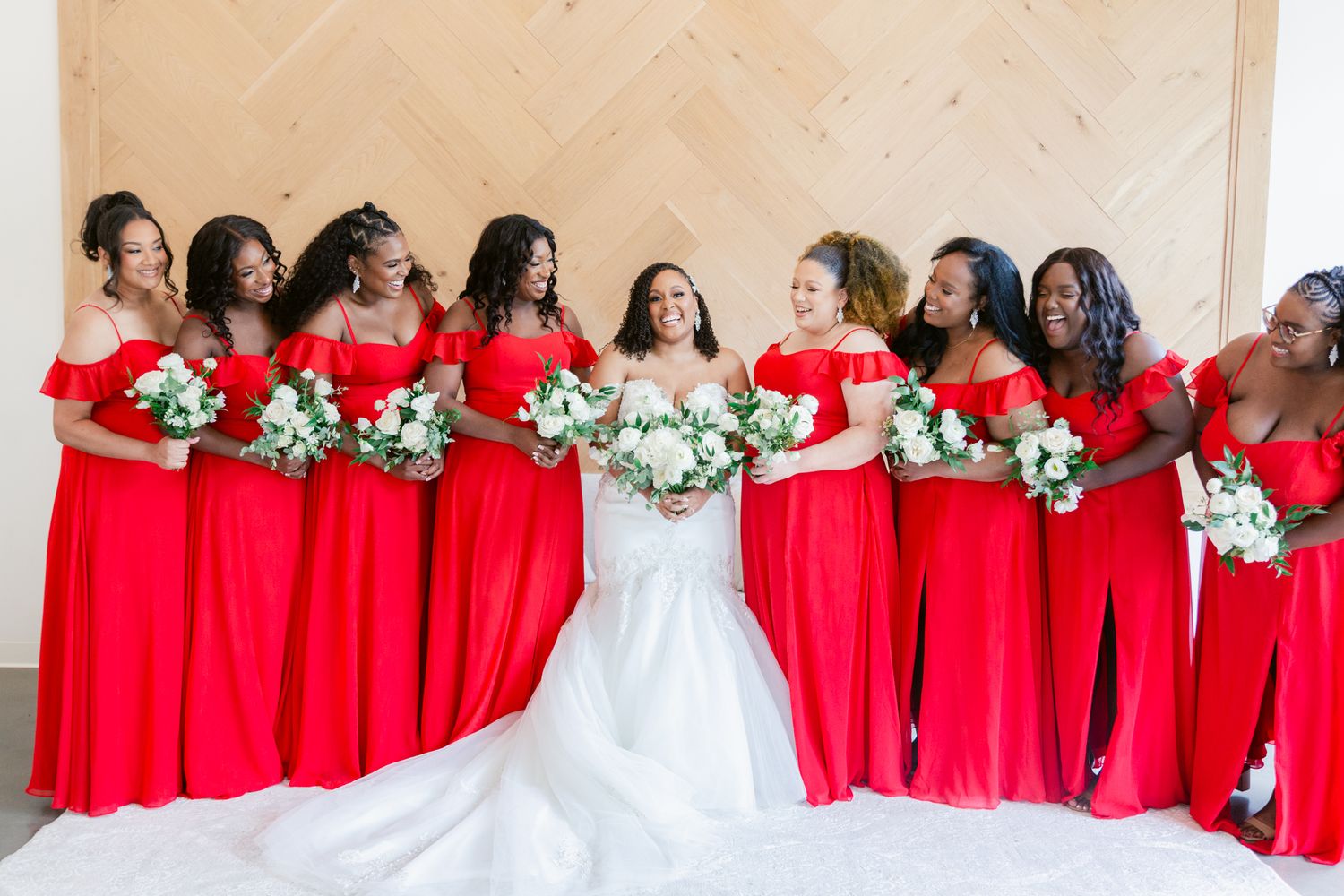 A wedding party in red bridesmaid dresses poses with the bride in white, holding white and green bouquets.