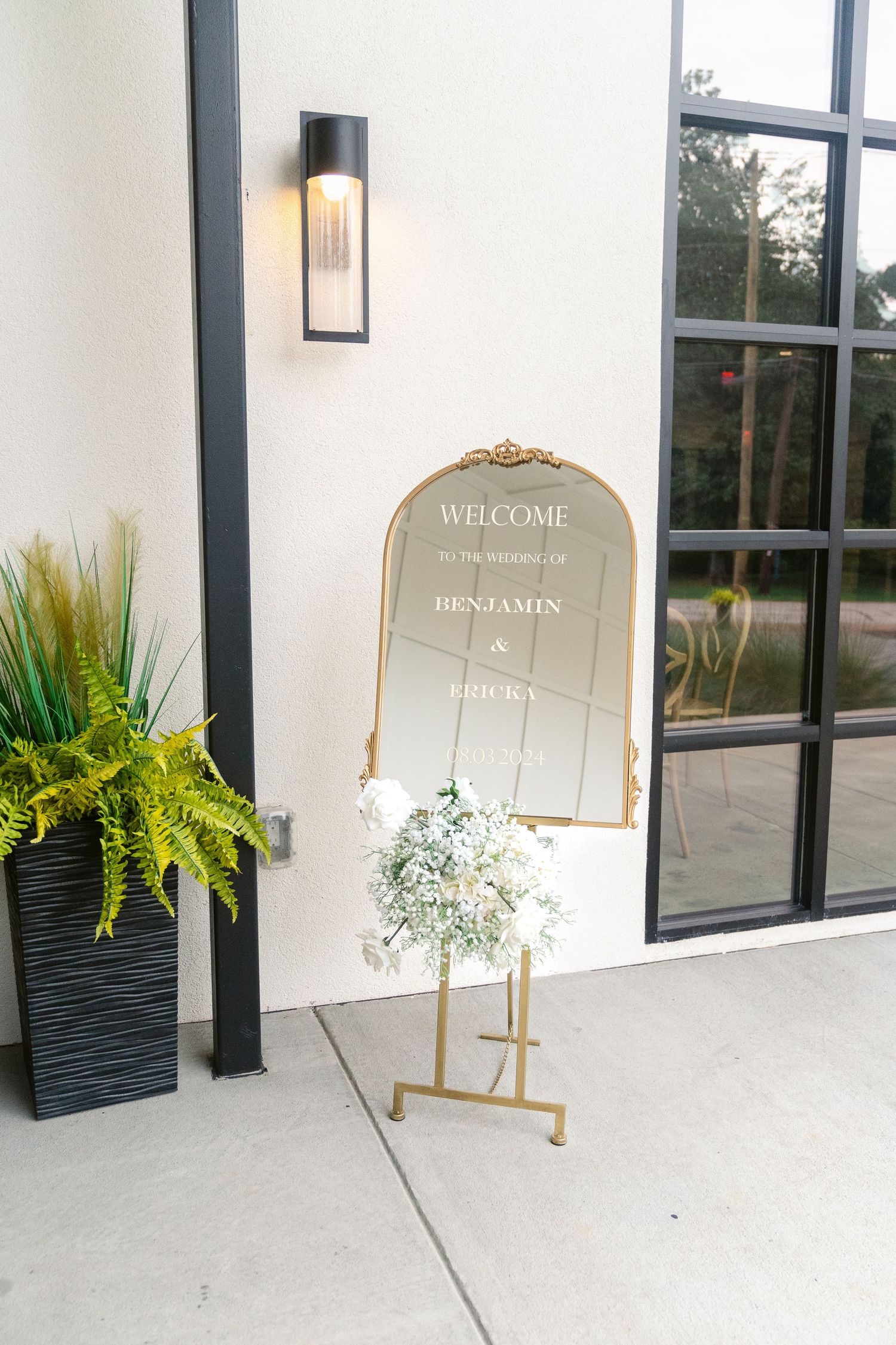Elegant gold-framed welcome mirror sign with potted fern decoration outside venue entrance.