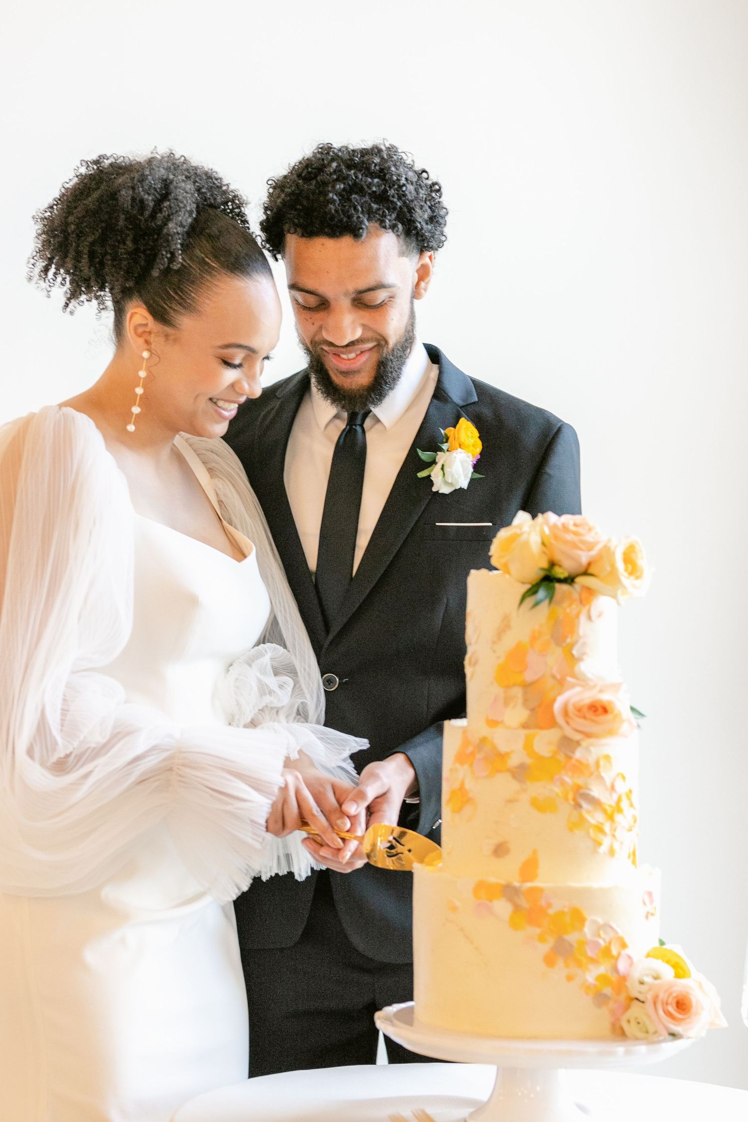 A couple in wedding attire cuts into an elegant peach-colored wedding cake adorned with floral decorations.