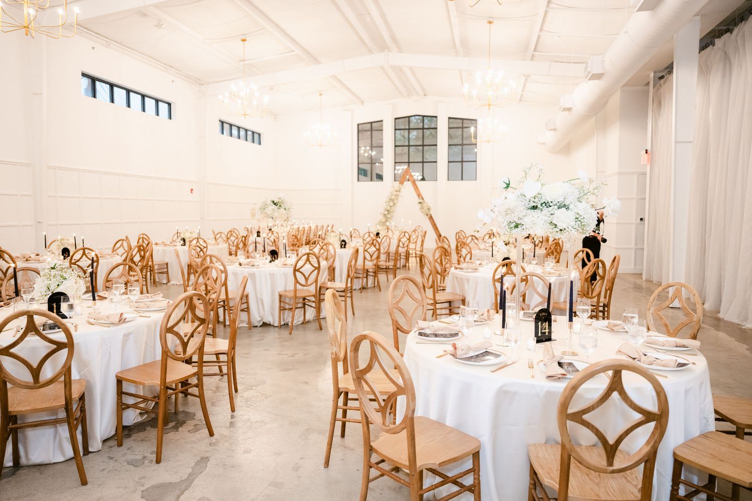Elegant white reception hall with wooden chairs and round tables set for a formal event.