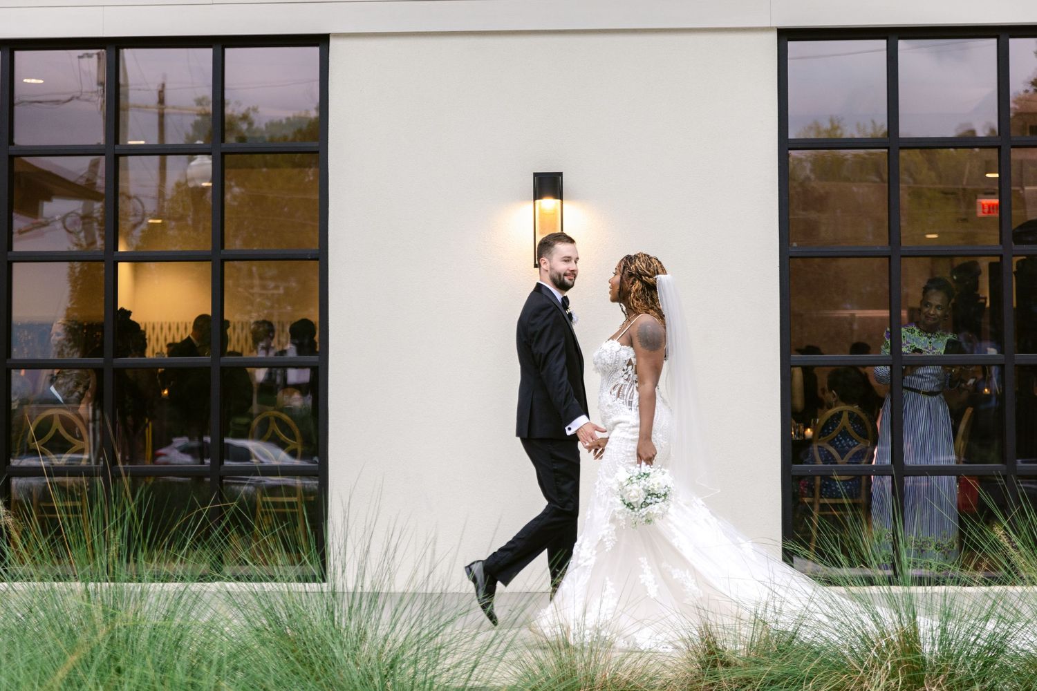 A wedding couple walks together along a modern building exterior with large windows at sunset.