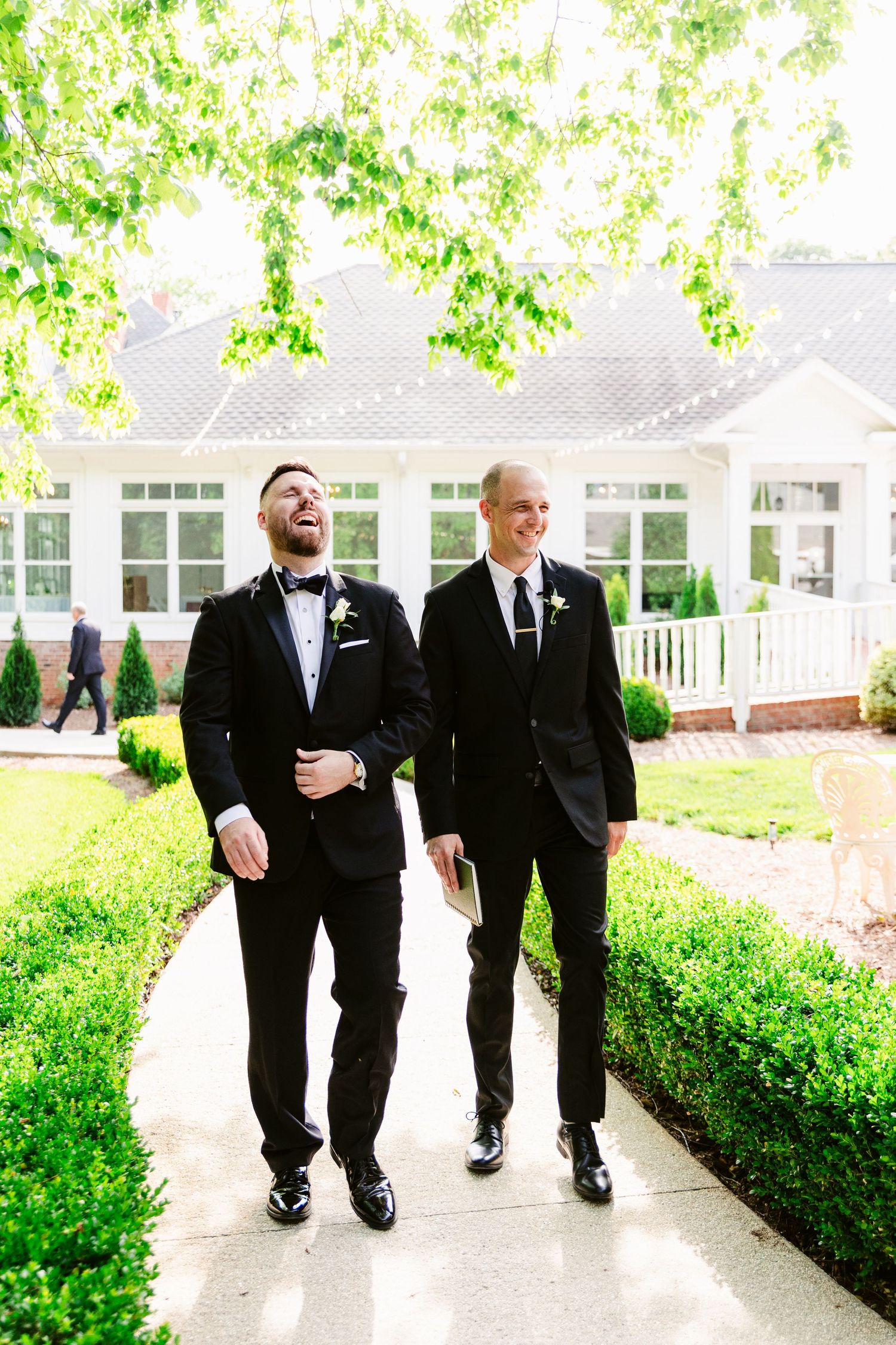 Four people in black suits walking and laughing together on a garden path.