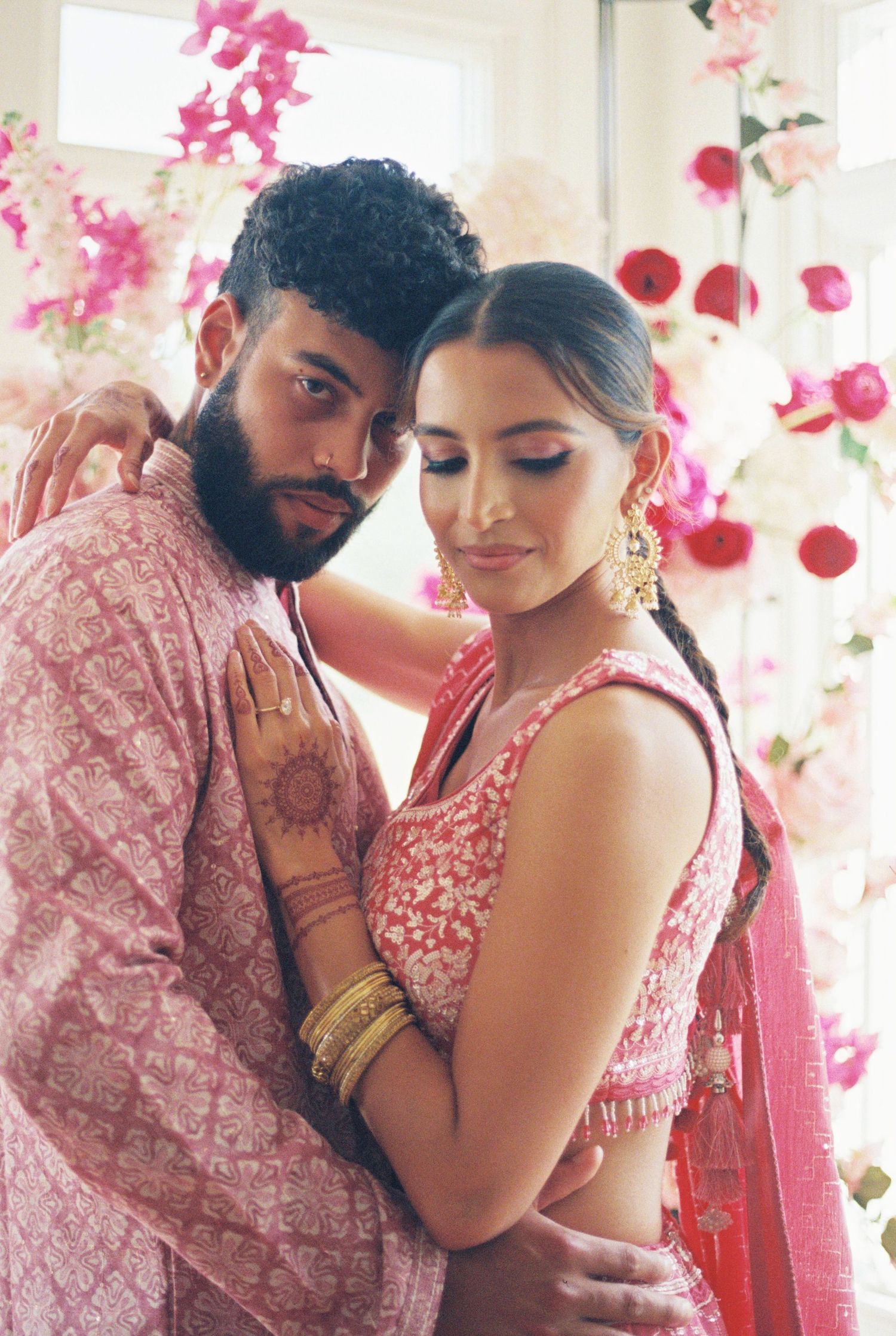 Romantic couple in traditional Indian wedding attire sharing intimate moments against pink floral backdrop.