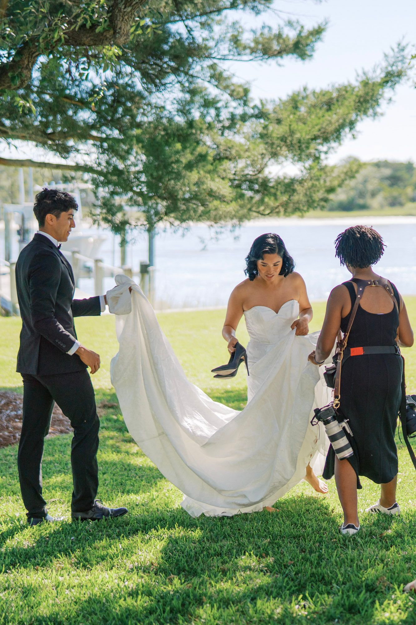 Wedding party helps carry a flowing white dress train across a grassy lawn by a waterfront on a sunny day.