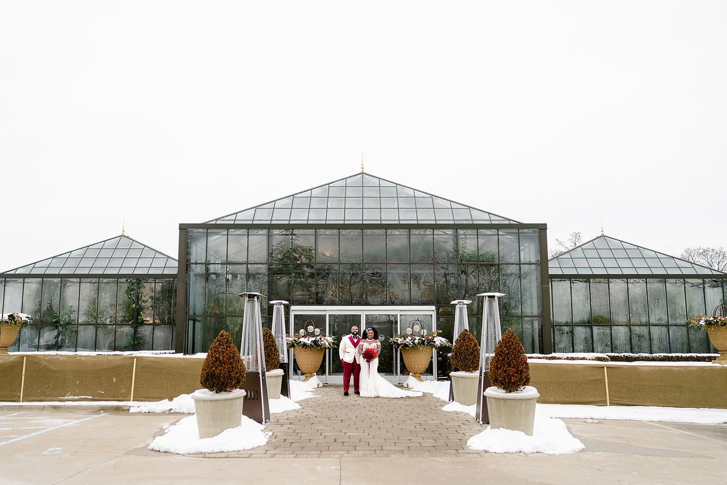 A modern glass greenhouse entrance with snow on the ground and people standing by the doors in winter.