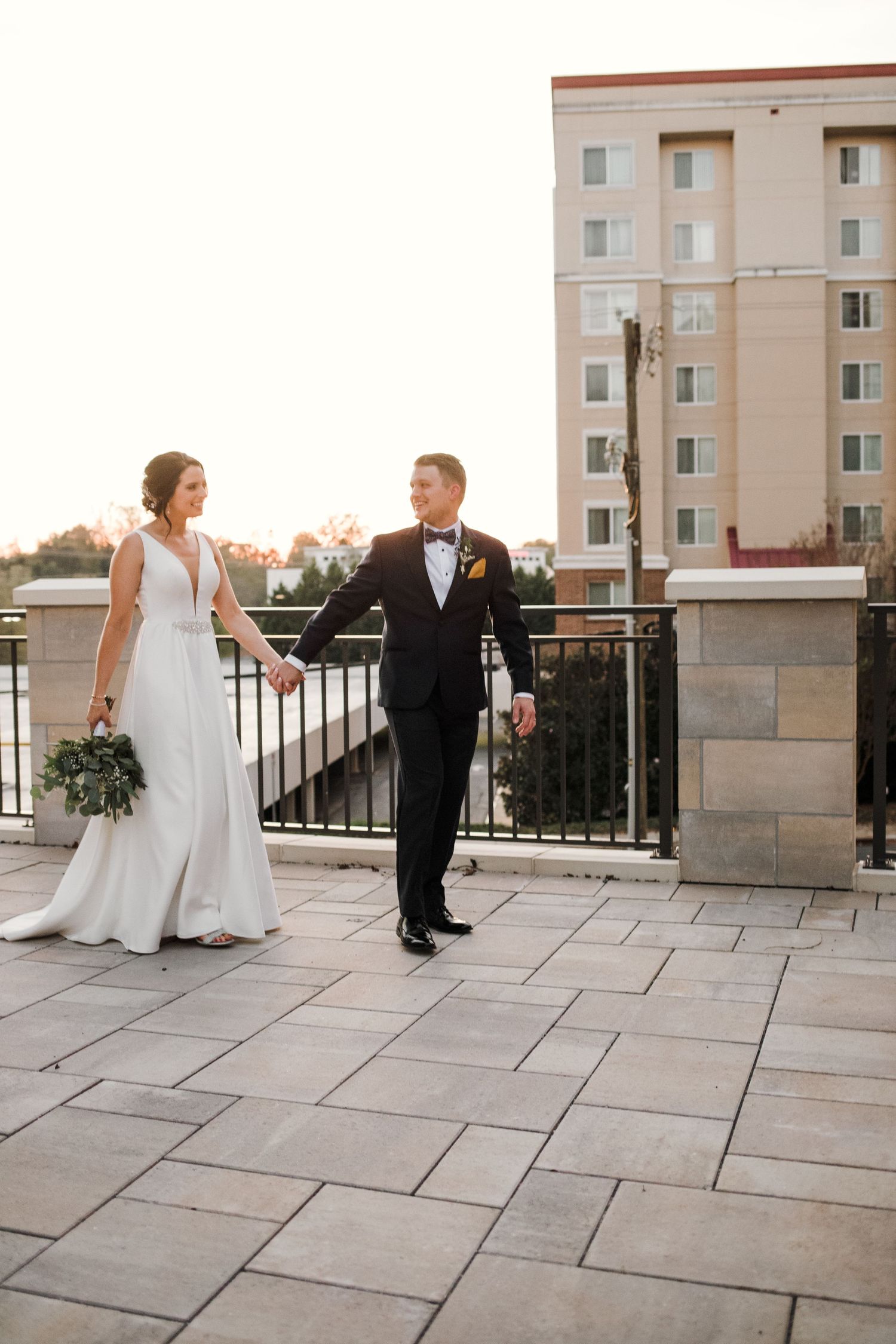 A newlywed couple walking hand in hand on a stone terrace with city buildings in the background.