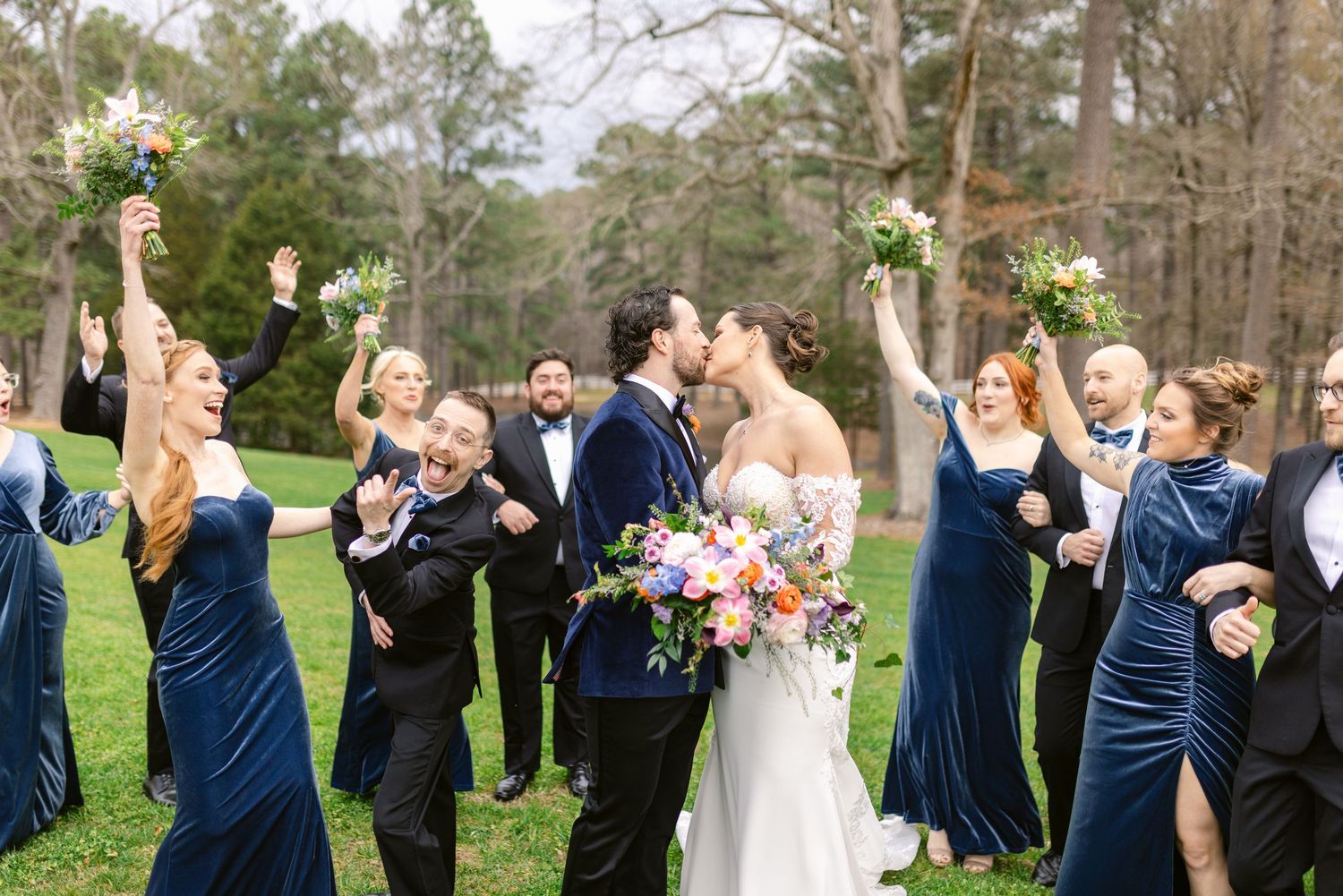Wedding party in navy blue attire celebrates with raised bouquets outdoors.