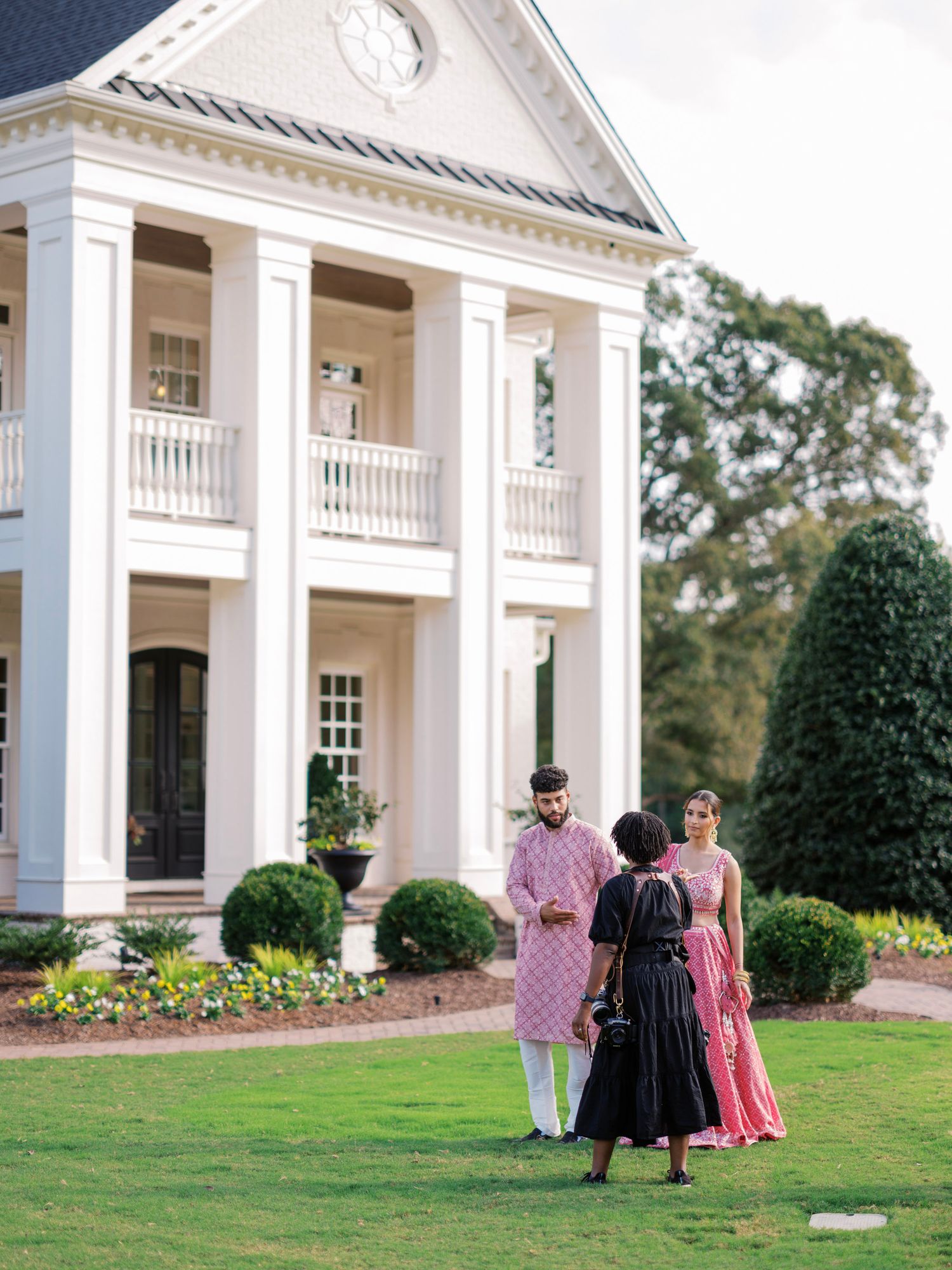 Elegant white colonial mansion with tall columns and manicured grounds serves as backdrop for outdoor gathering.