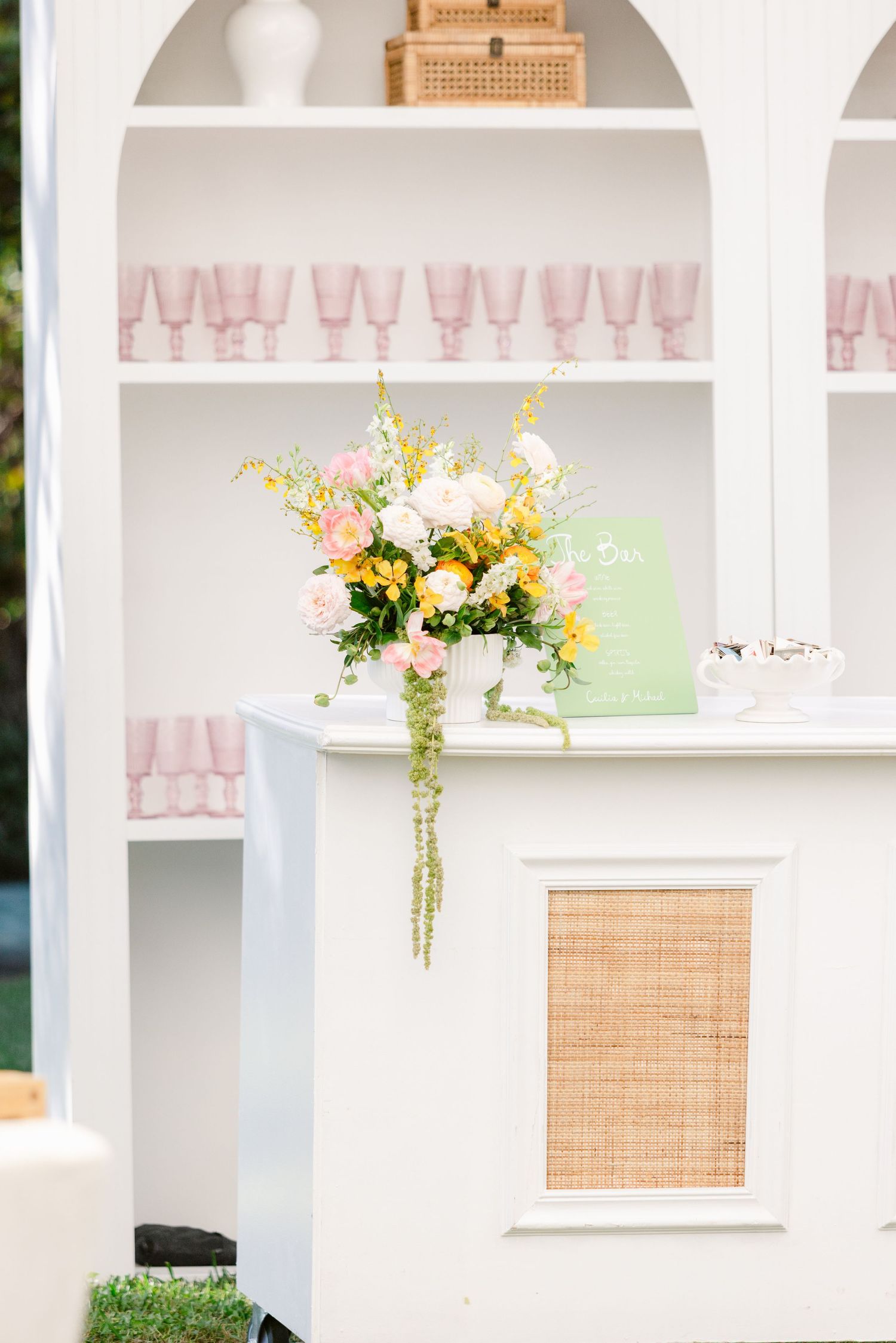 Elegant white bar display with pink glassware and floral arrangement featuring white and yellow blooms.
