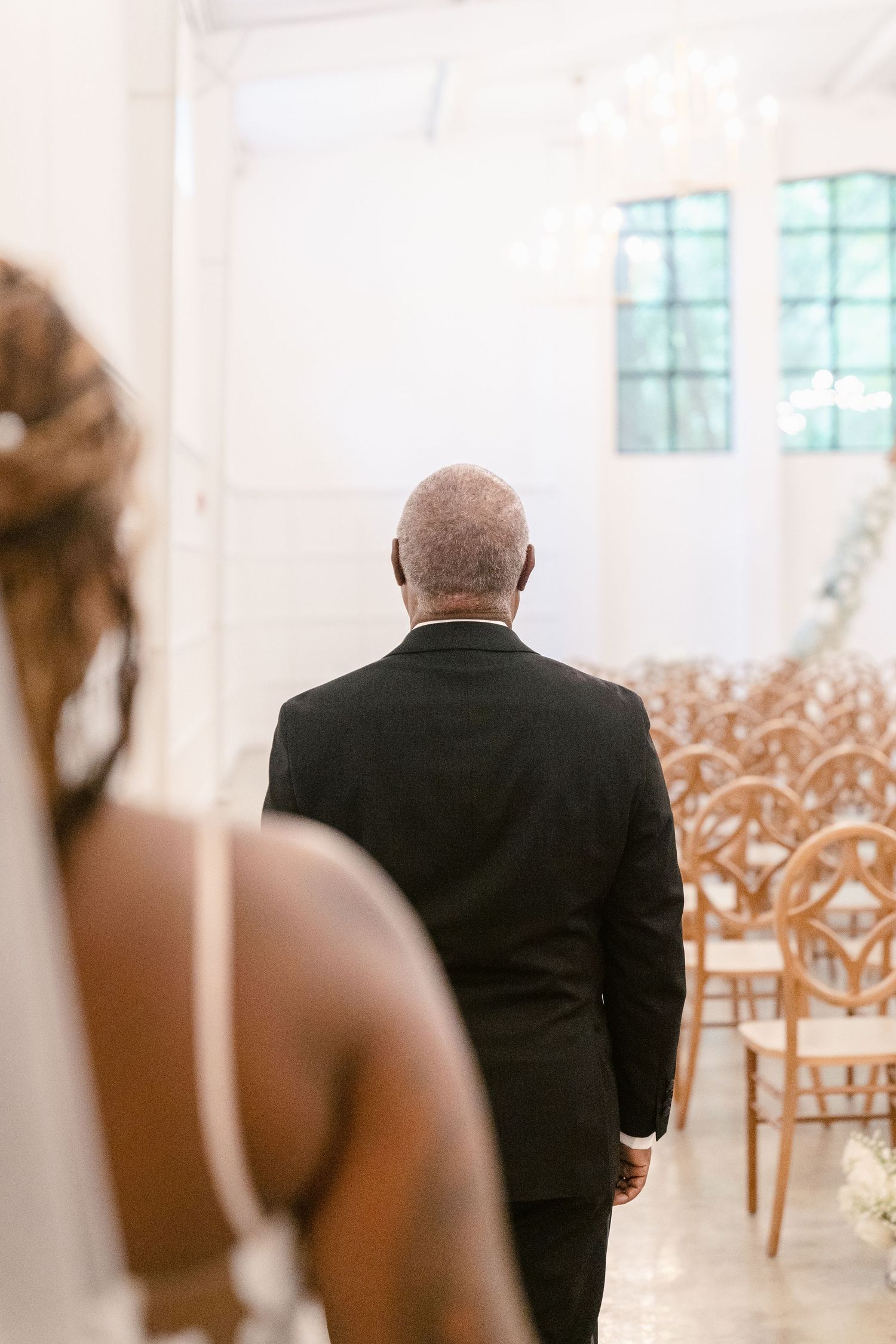 An elderly man in a black suit walks away down a bright white aisle lined with ornate chairs at a wedding venue.