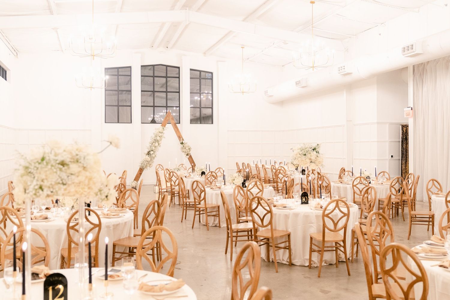 Light-filled wedding reception venue with wooden chairs and white tablecloths set for an elegant celebration.