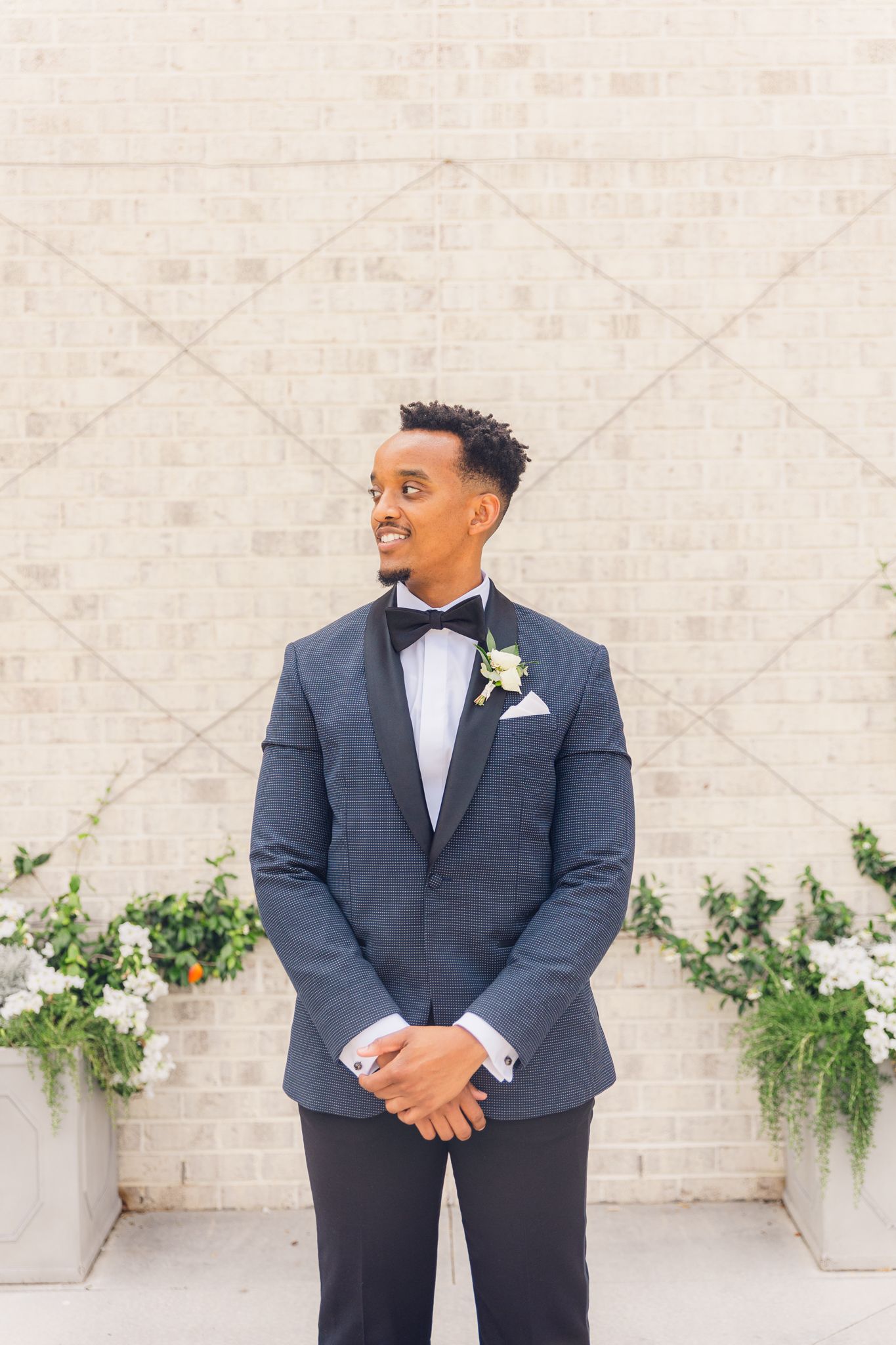 Groom in navy tuxedo poses with joyful expression against white wall with decorative greenery.