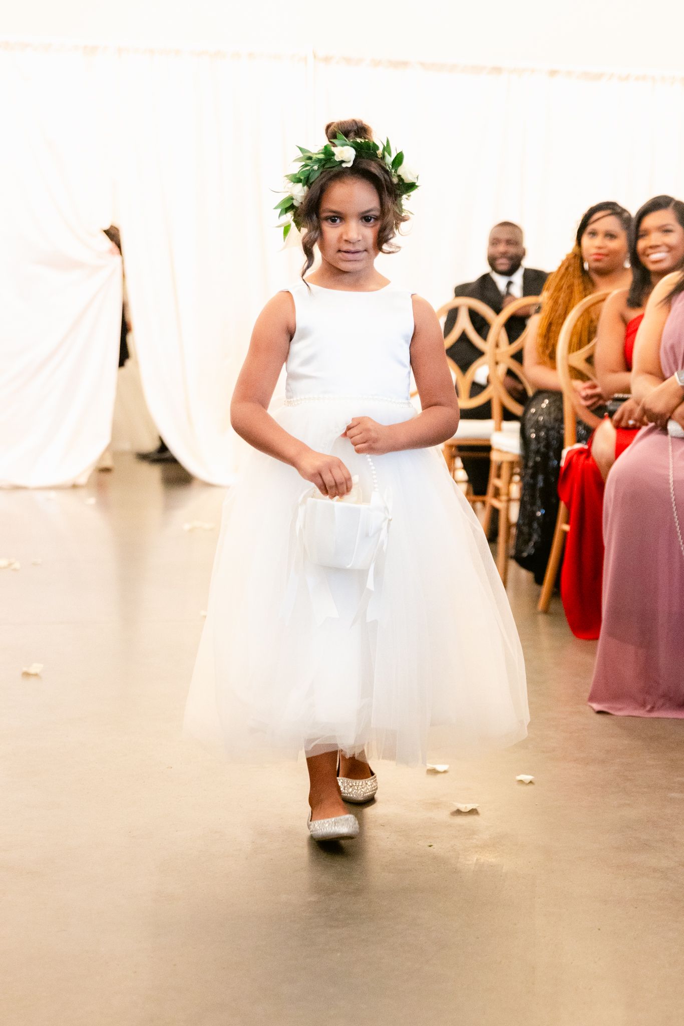 A flower girl in a white dress walks down the aisle at an elegant wedding ceremony with guests seated on both sides.