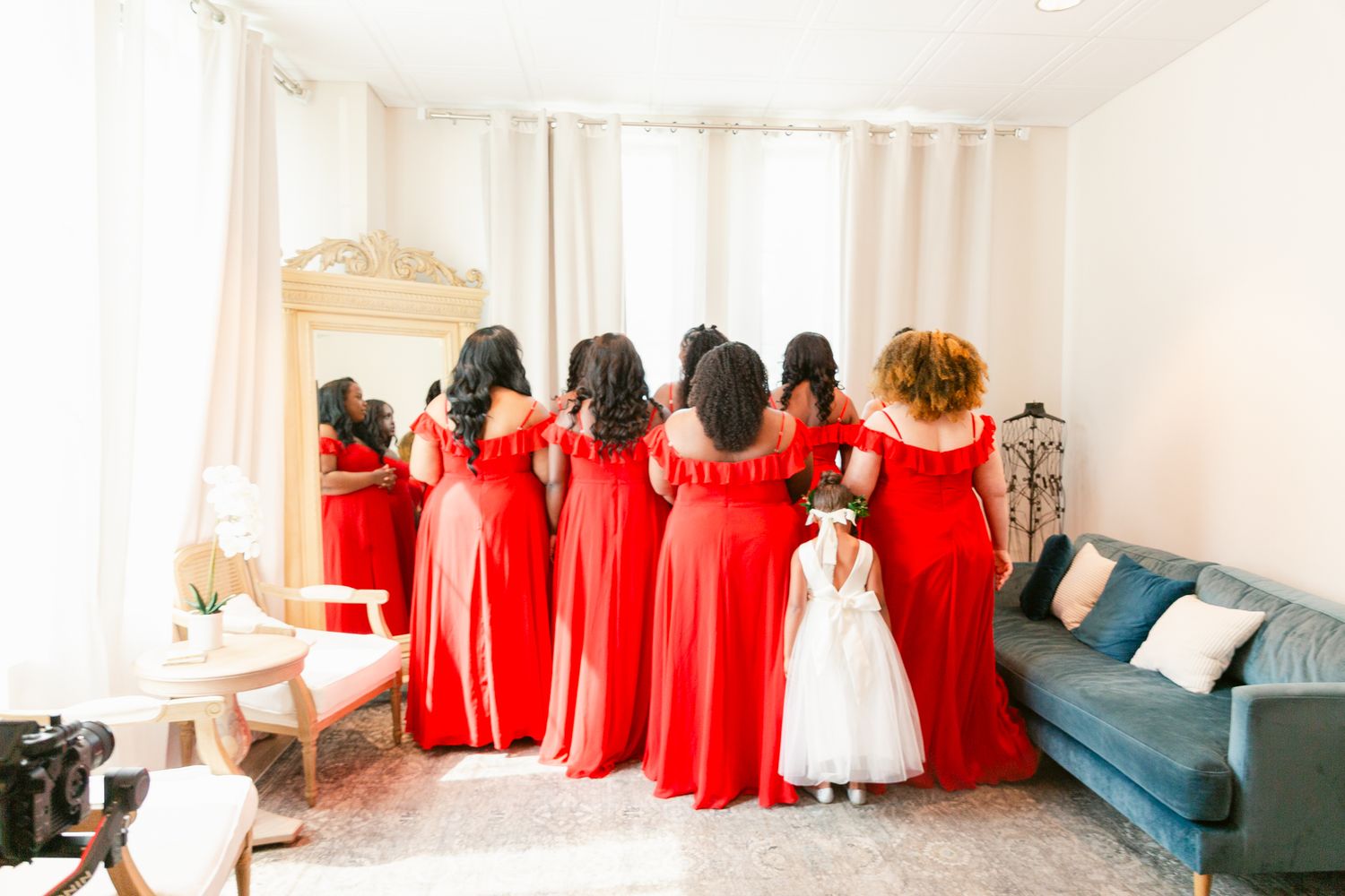 Bridesmaids wearing matching long red dresses stand together with a young flower girl in white in a bright room.