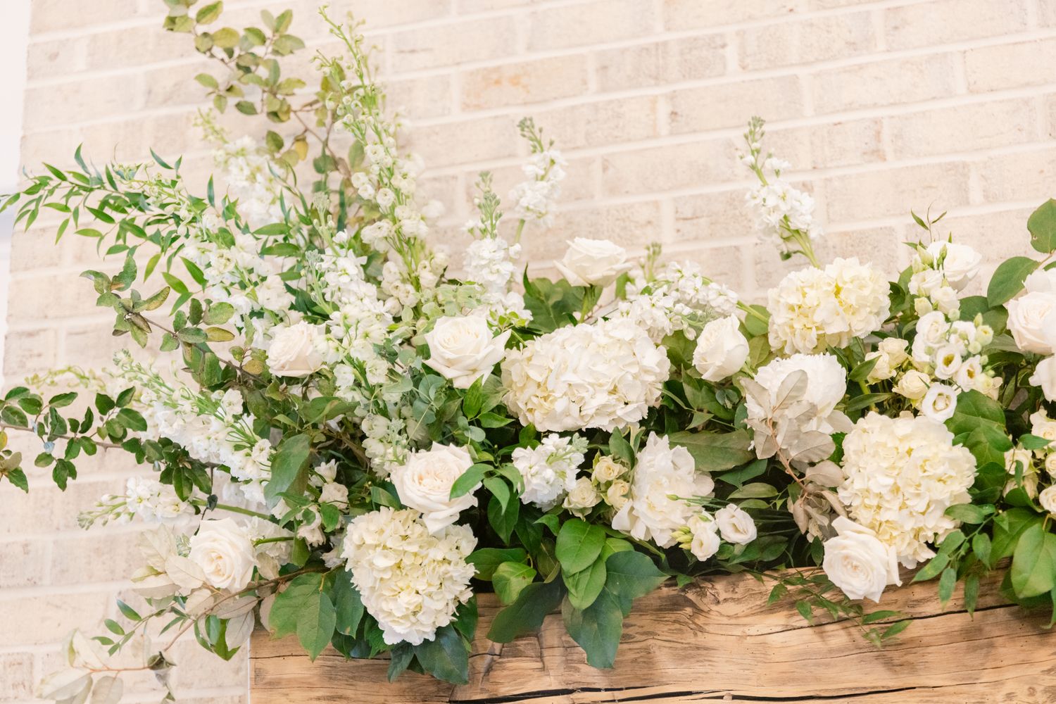 Lush floral arrangement featuring white hydrangeas and roses with greenery against a brick wall.