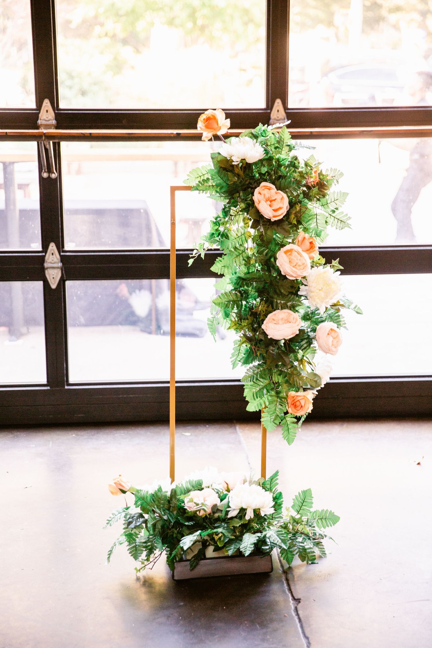 Modern floral arrangement with peach roses and greenery cascading down a gold metal frame stand by a window.