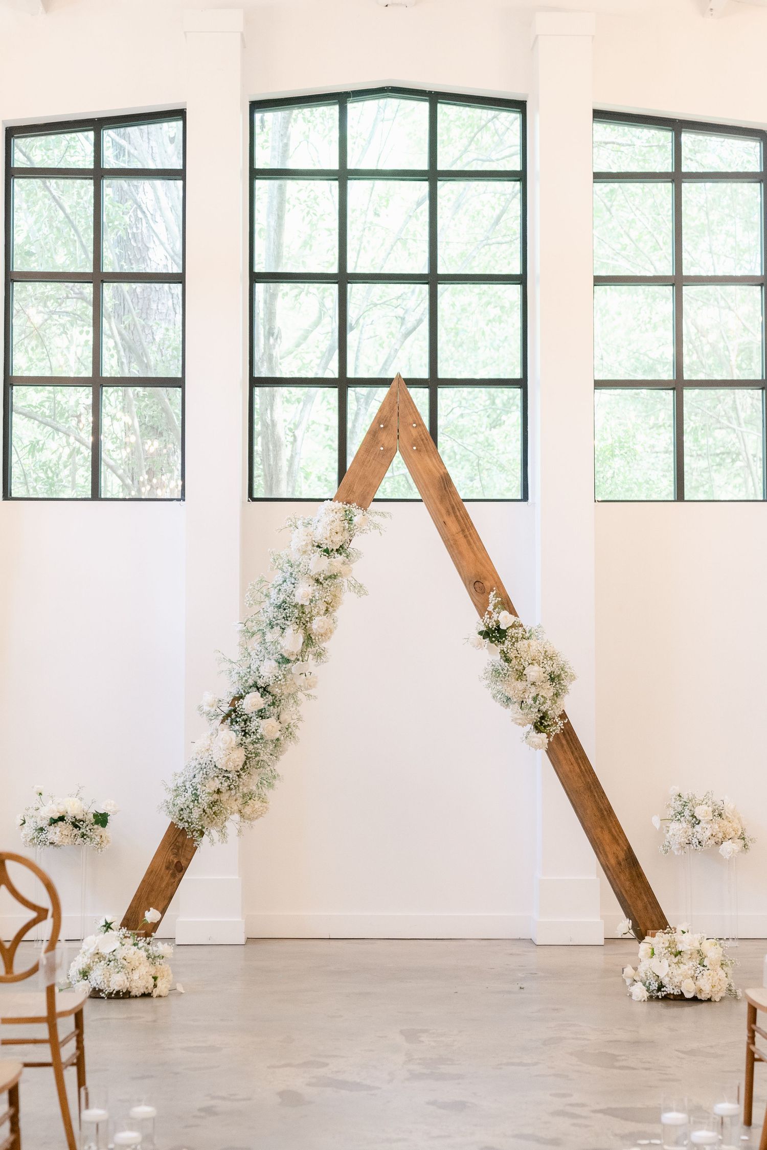 Modern wedding ceremony setup with wooden triangle arches and white floral arrangements in an industrial space.