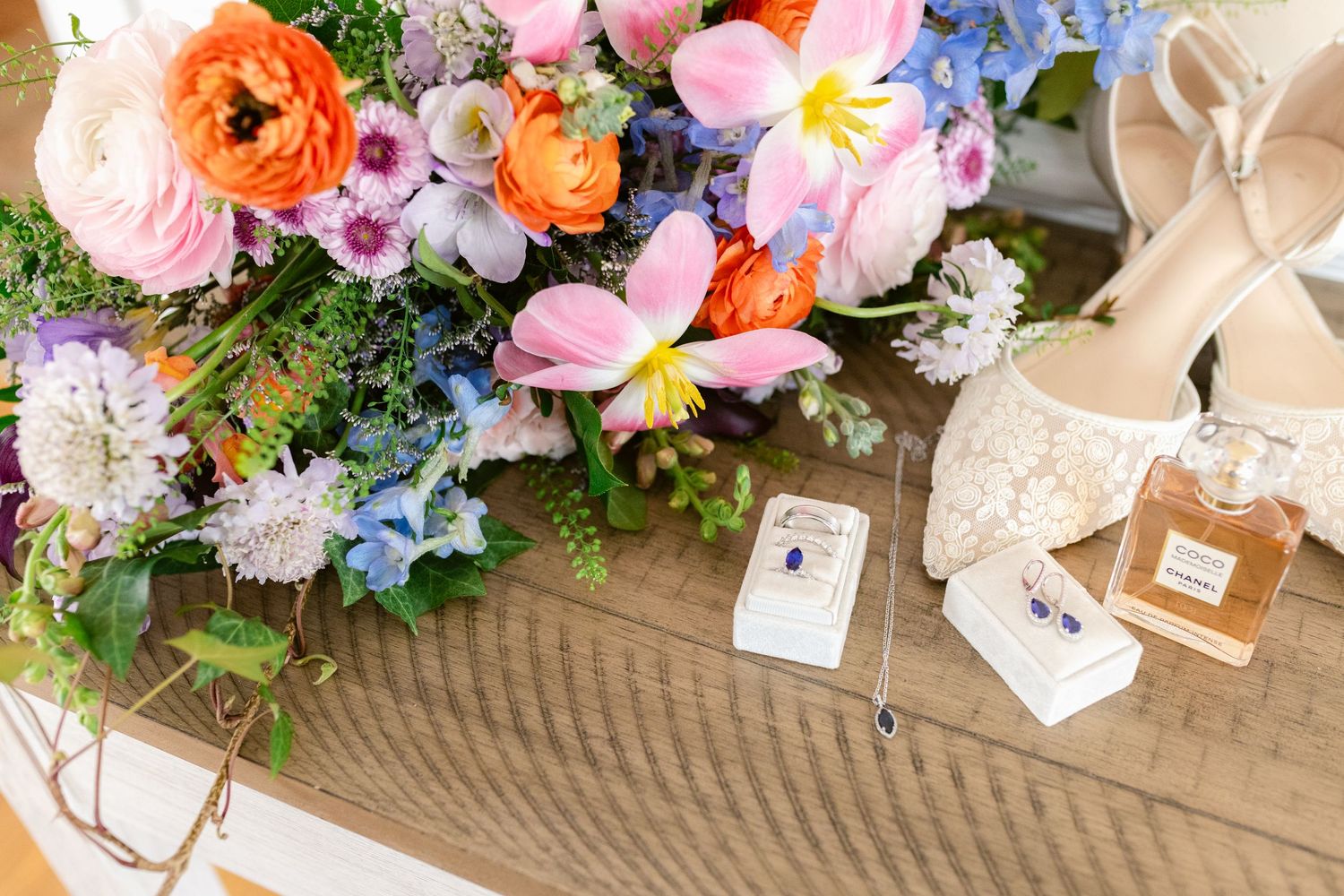 Wedding rings and perfume bottles beside colorful spring flowers including ranunculus and orchids on wooden surface.