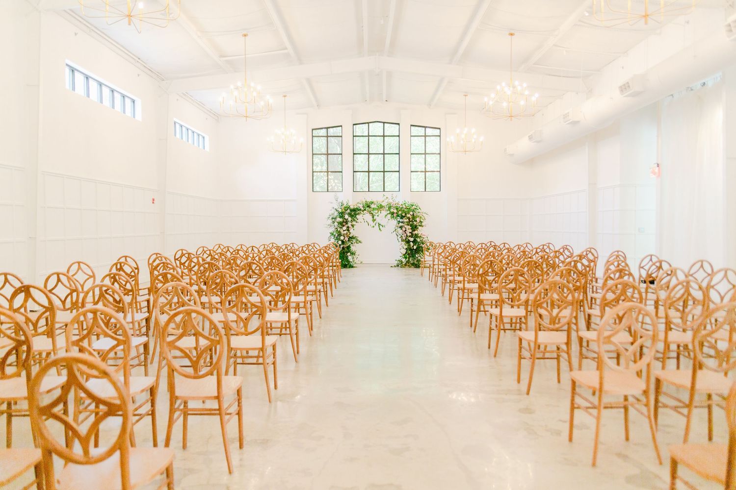 Light-filled event space with rows of wooden chairs arranged in a ceremony setup under a vaulted white ceiling.