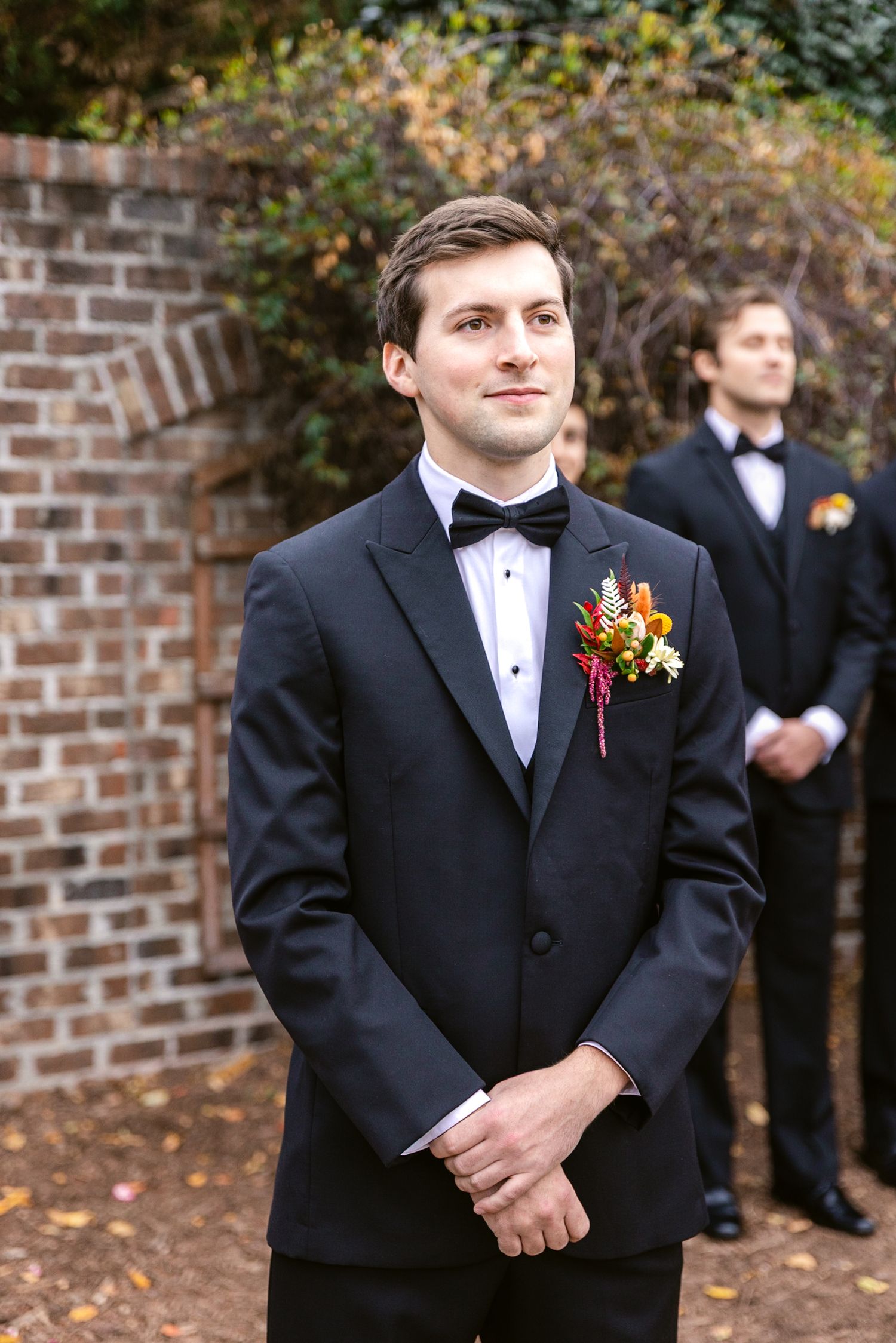 Groomsman in black tuxedo with autumn-colored boutonniere stands against brick wall at outdoor wedding ceremony.