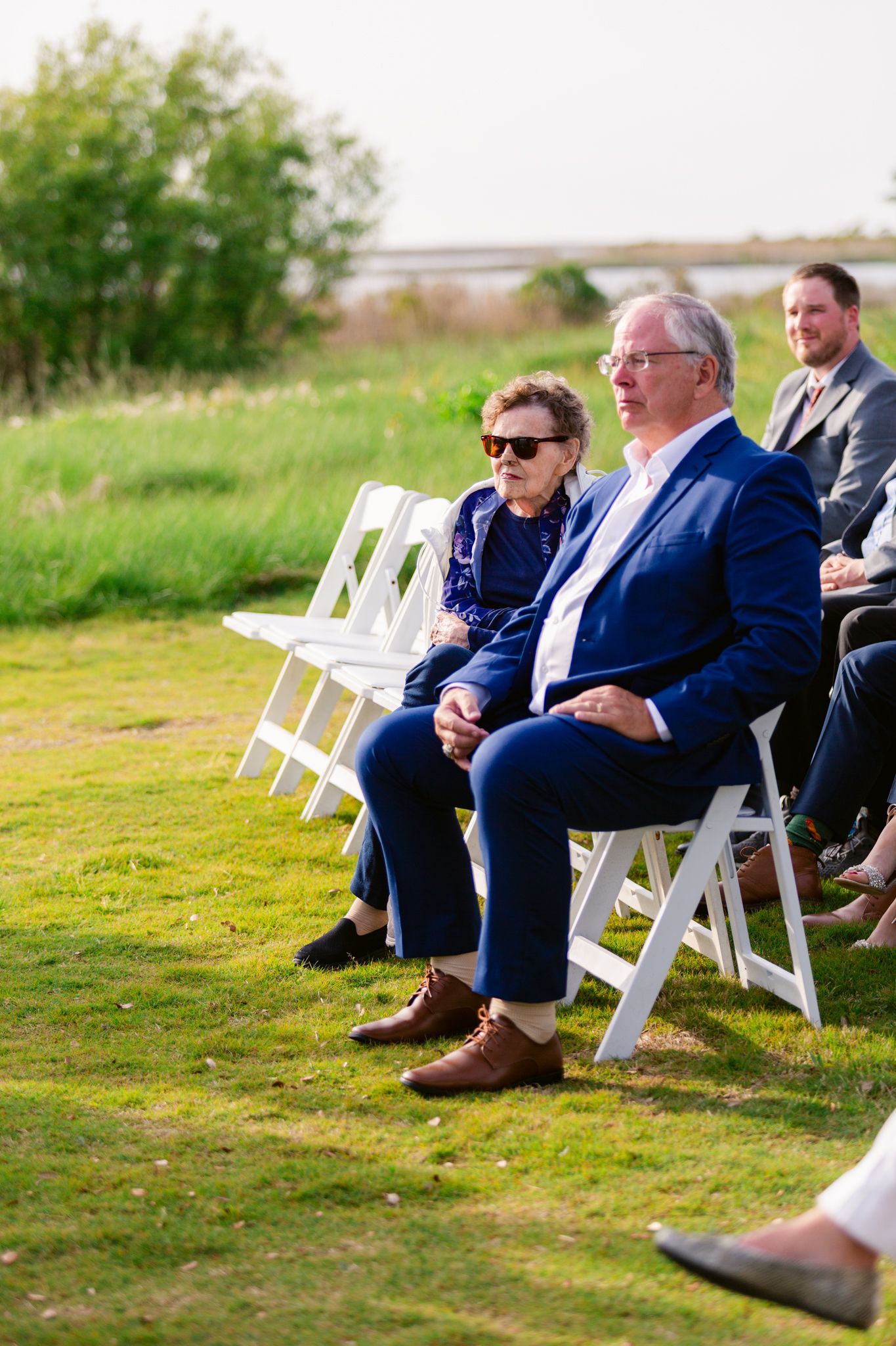 Wedding guests in navy suits sit on white folding chairs on a grassy lawn during an outdoor ceremony.