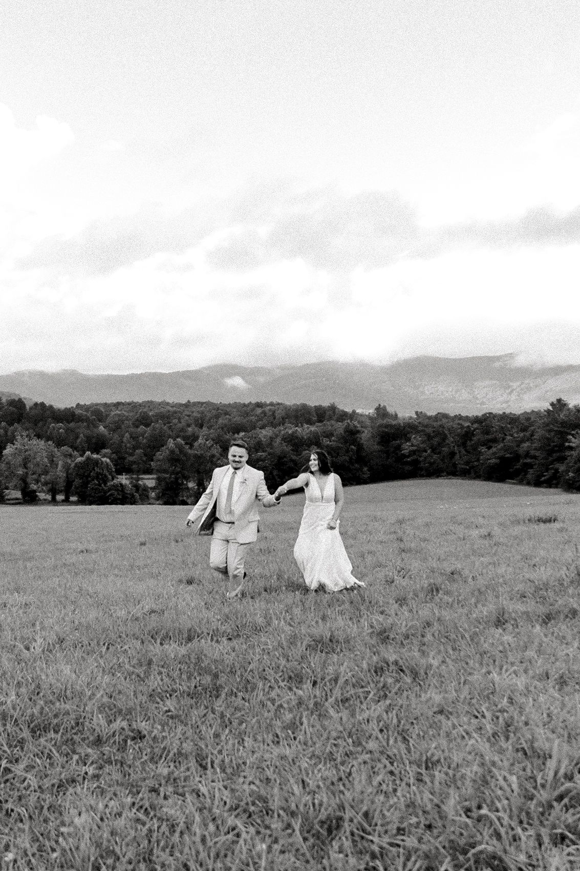 A couple in wedding attire walks through a scenic meadow in black and white photography.