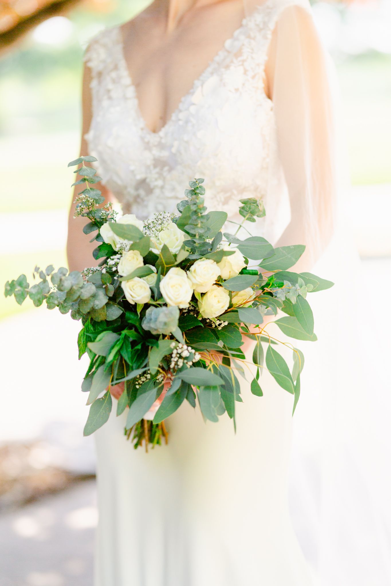 A delicate bridal bouquet featuring white roses and mixed eucalyptus greenery held against a white wedding dress.