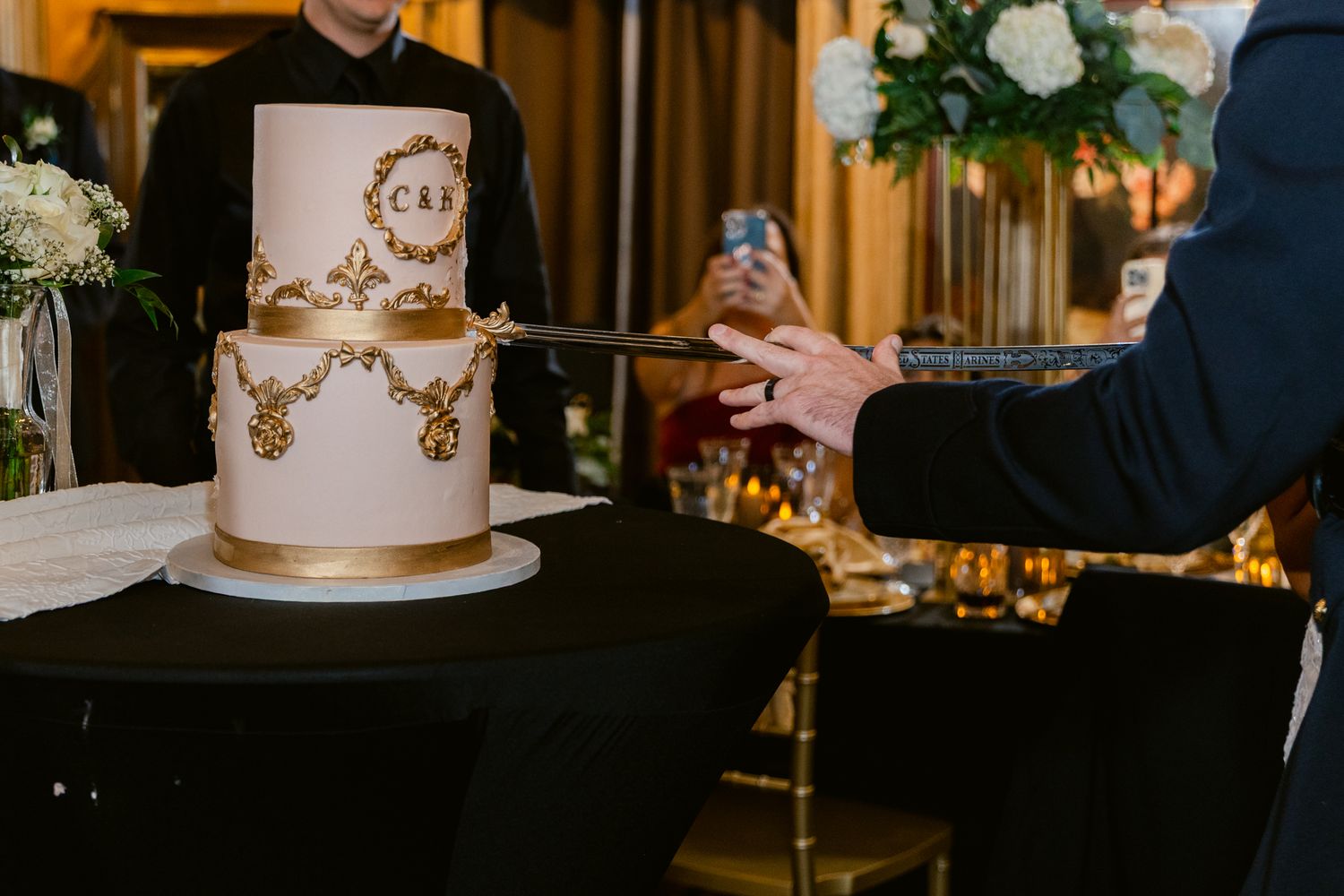 Elegant white and gold wedding cake with ornate metallic details displayed on black table at reception venue.