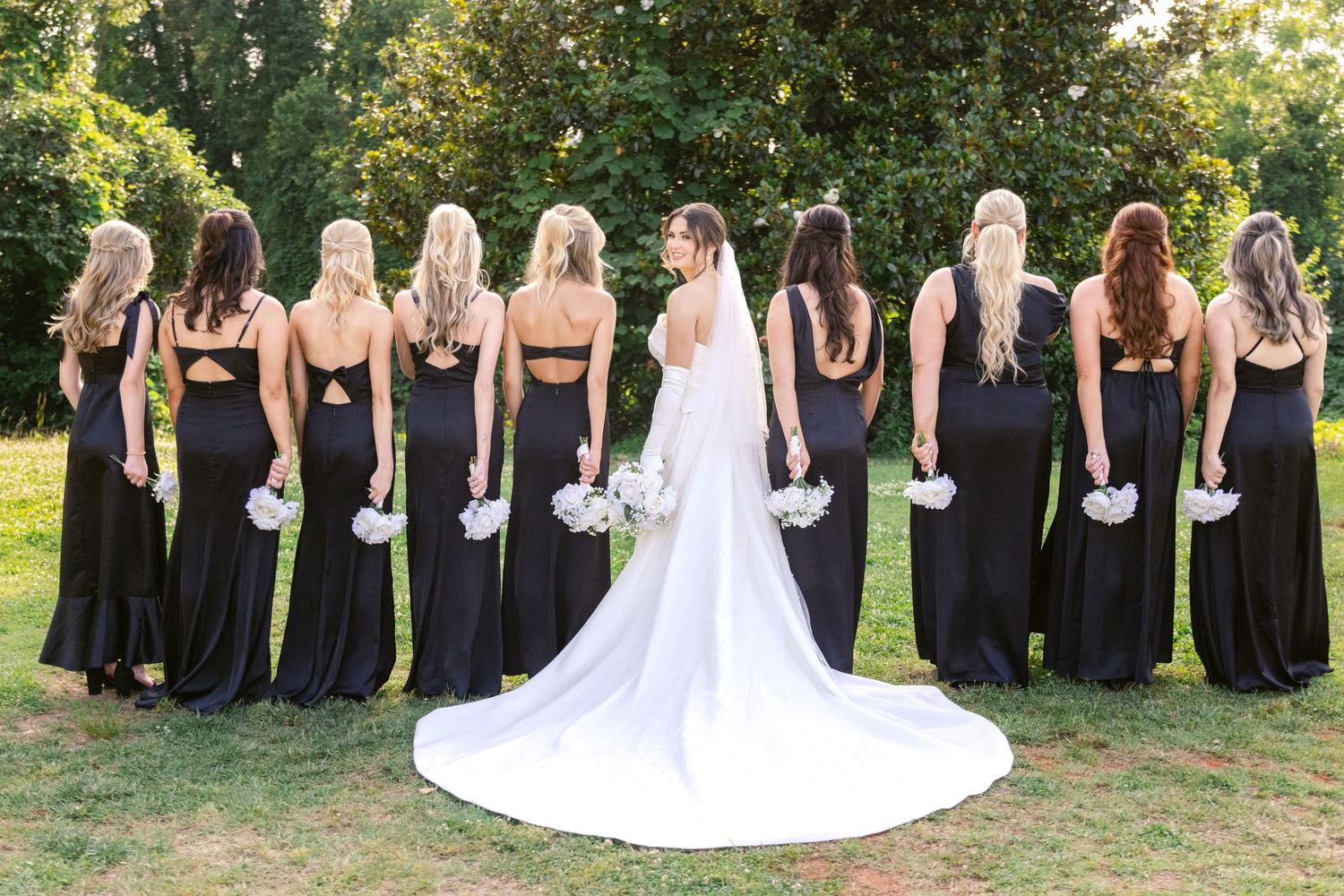 Wedding party in black bridesmaid dresses stands outdoors with bride in flowing white gown and long train.